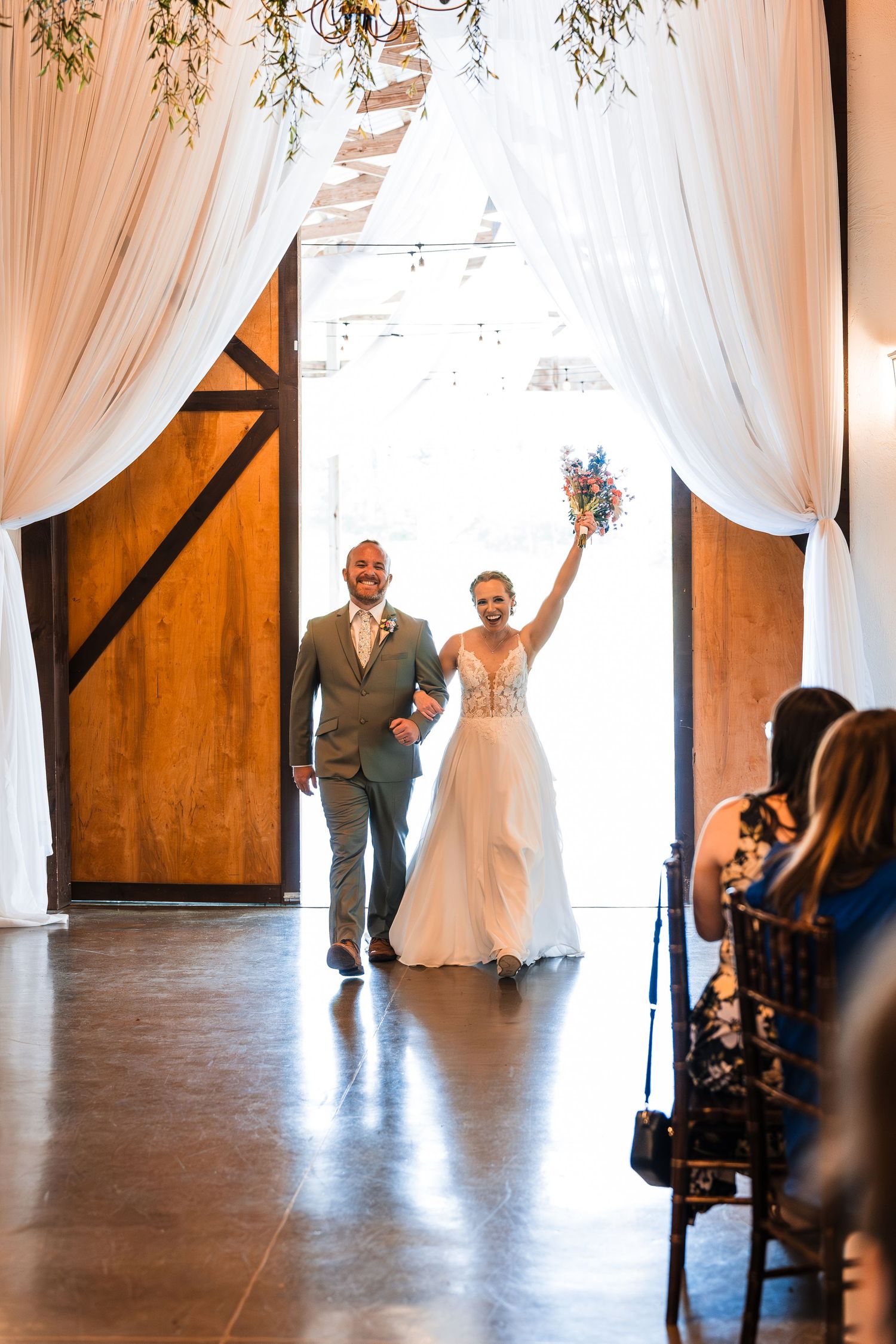 Bride raises bouquet triumphantly as couple enters barn wedding venue through draped white curtain doorway.