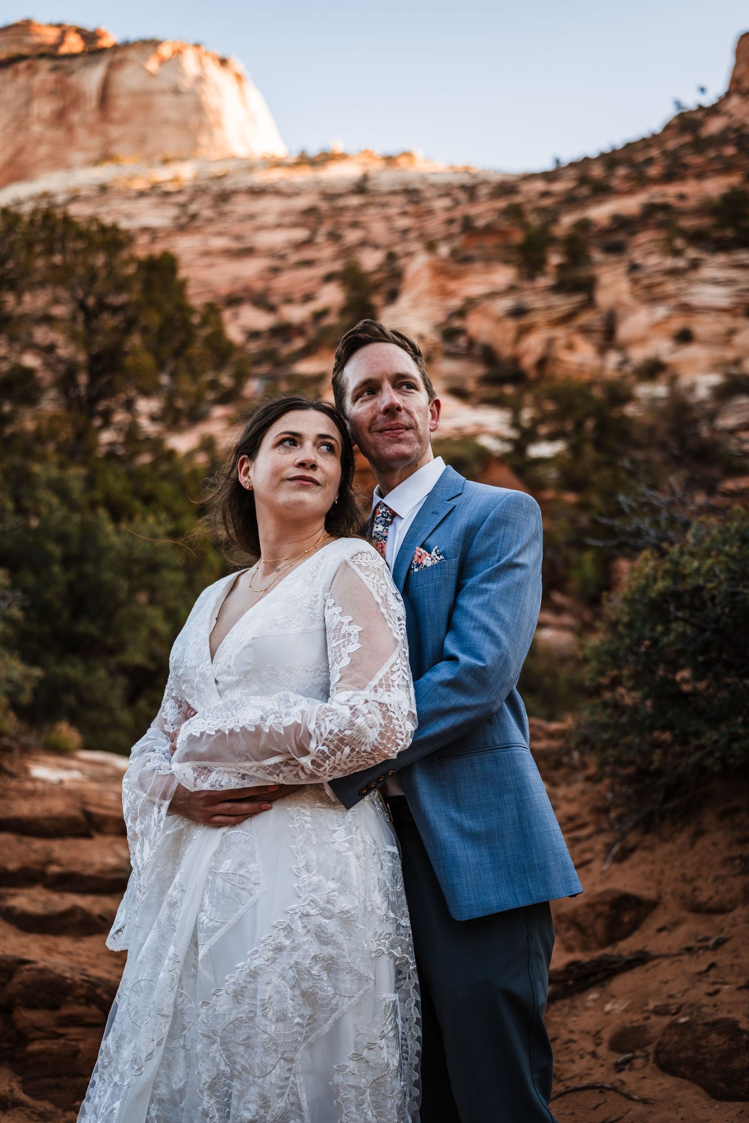 Bride and groom embrace while gazing upward against red sandstone cliffs at Zion National Park.