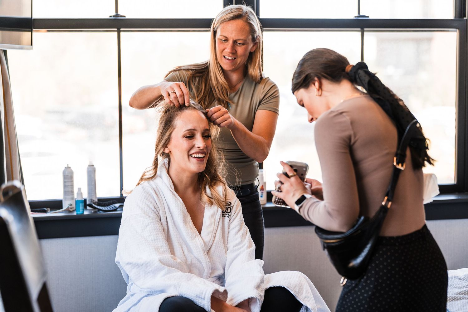 Bride in white robe laughing as two stylists do her hair and makeup before her wedding day.
