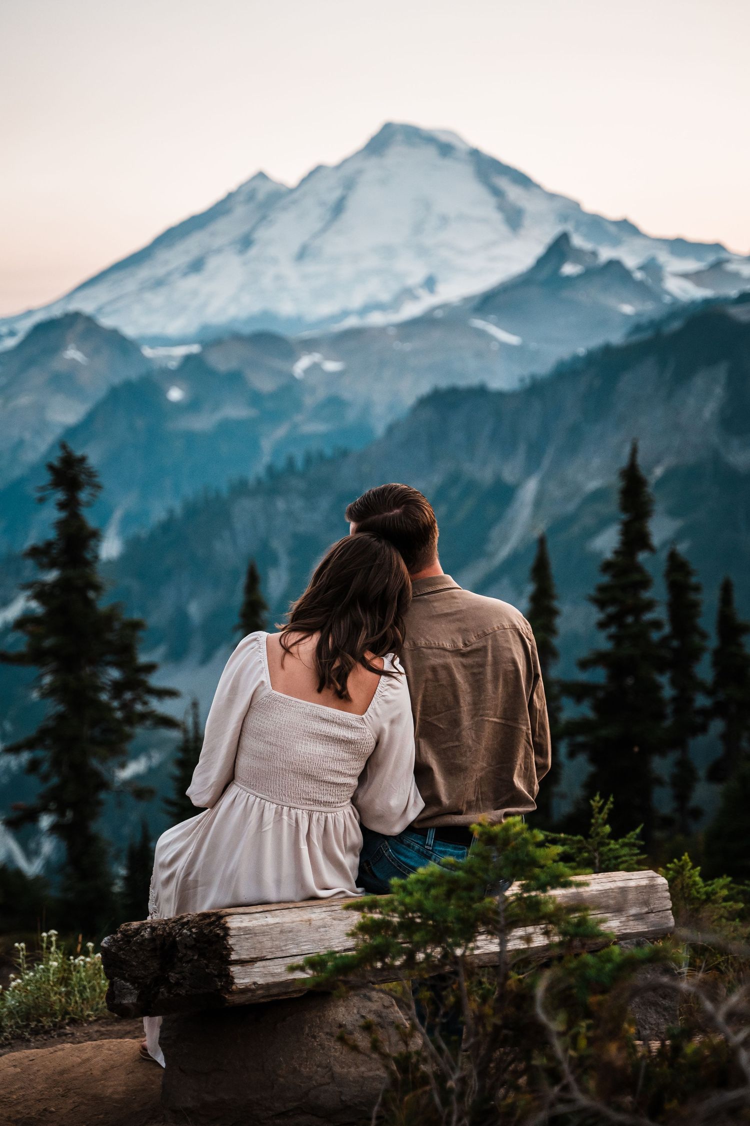 Two people sit together on a log overlooking majestic snow-capped mountain peaks and evergreen forest.