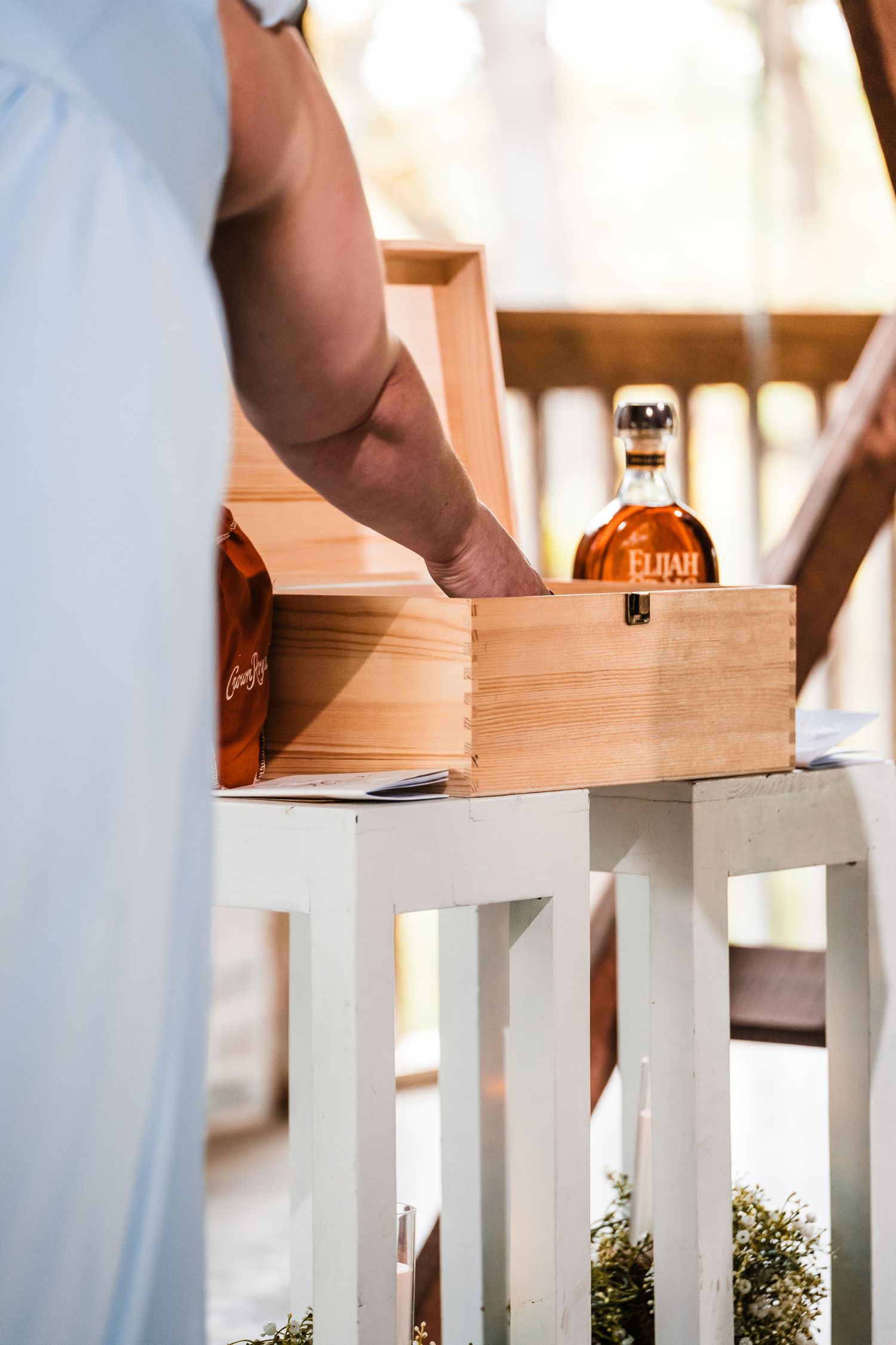 Hand reaches for a bottle in a wooden box displayed on white-painted wood railing during wedding ceremony setup.
