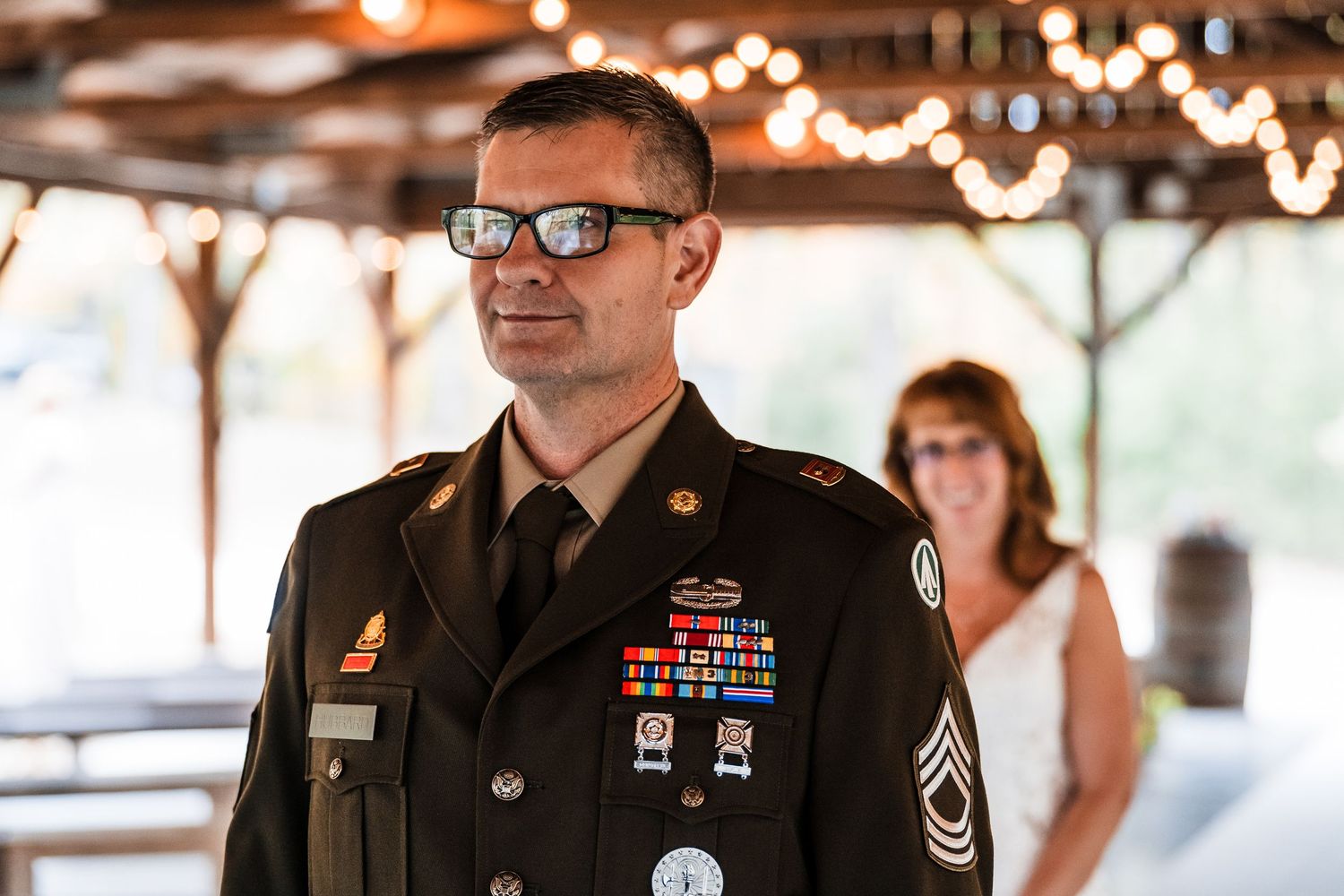 Military service members stand in formal dress uniforms at an indoor event with string lights overhead.