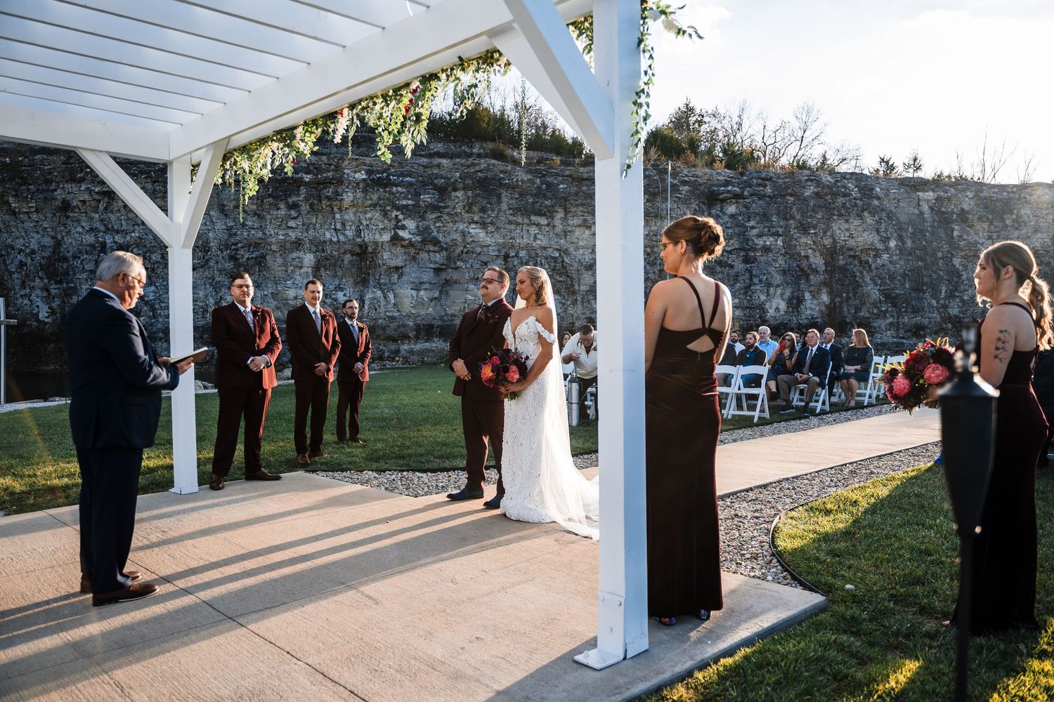 Wedding ceremony setup under a white pergola with guests gathering at sunset.