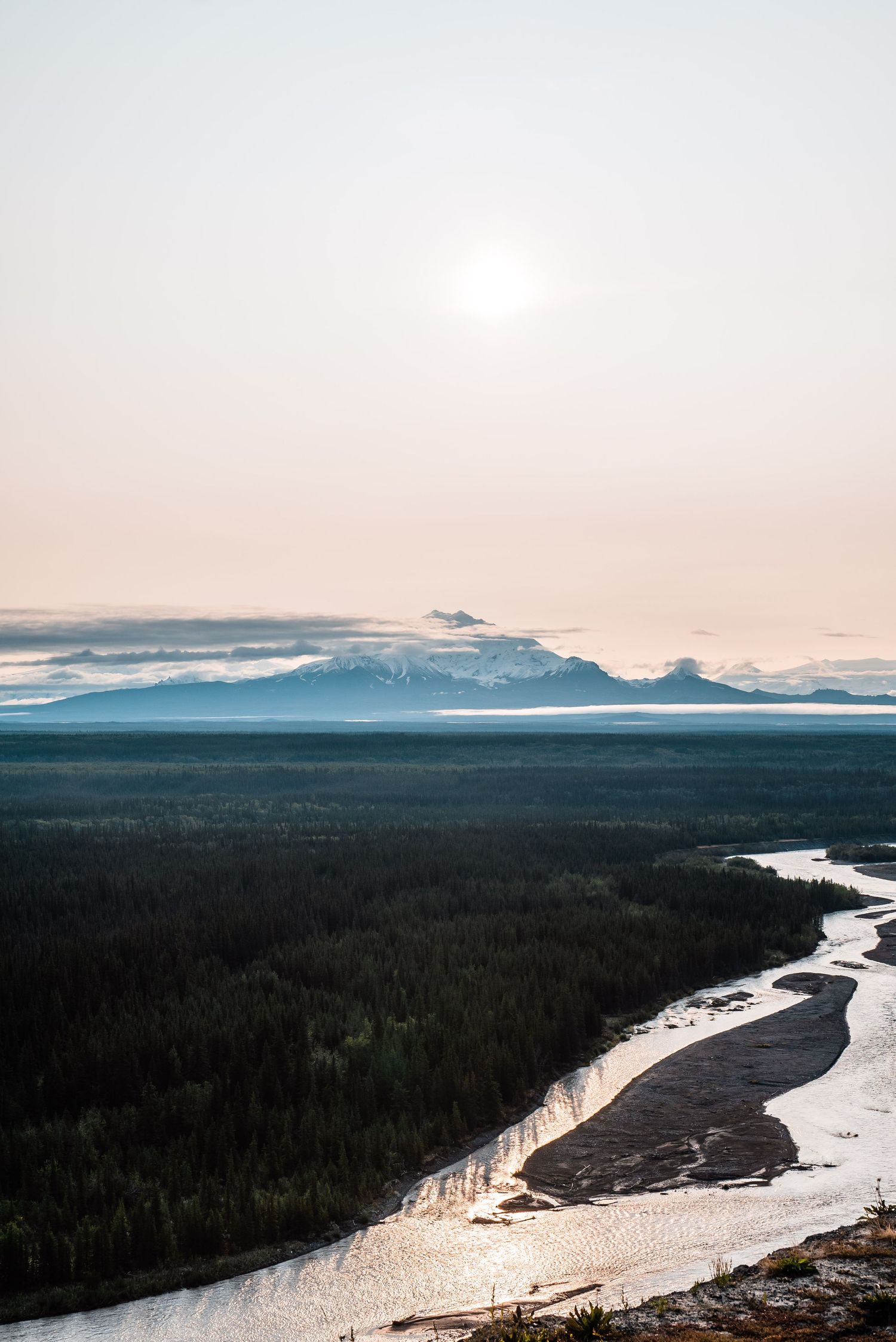 A winding river reflects sunset light through a forested valley with snow-capped mountains in the distance.