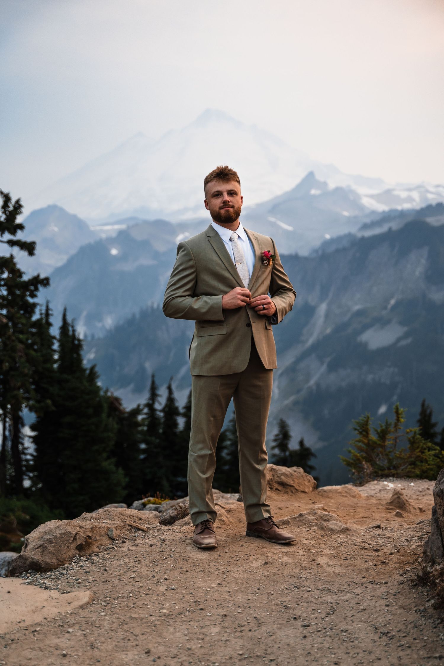 A person in a gray suit stands on a rocky mountain overlook with snow-capped peaks in the distance.