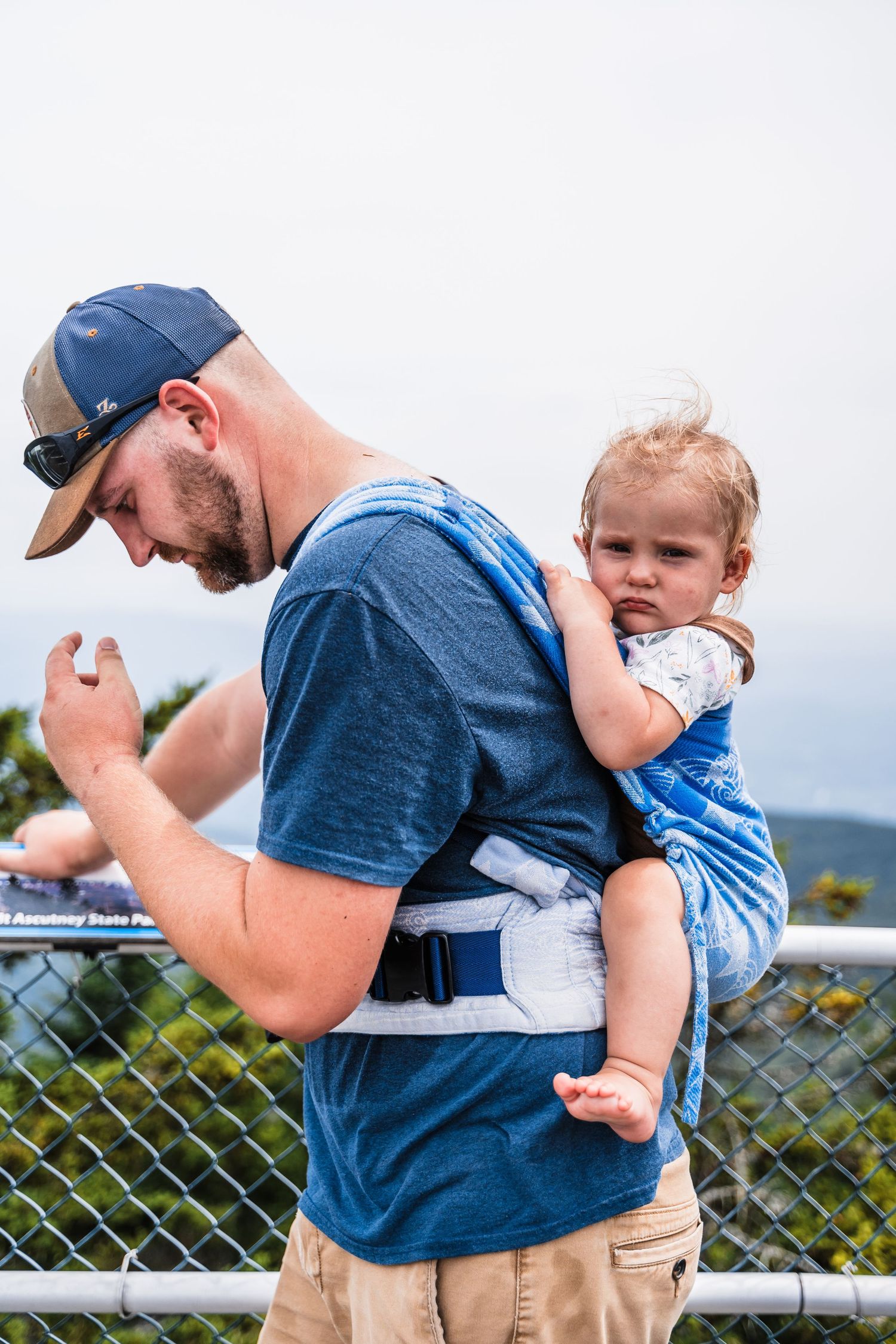 Parent carries young child in a blue baby carrier while standing near a scenic overlook.