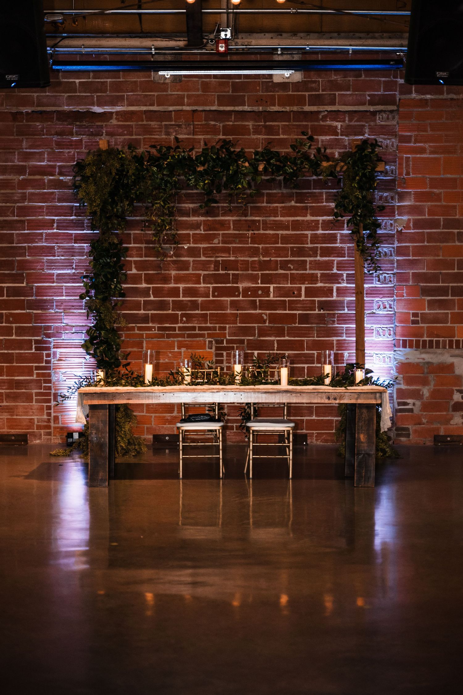 Wedding ceremony setup with greenery arch and rustic wooden table against exposed brick wall lit by soft purple lighting.