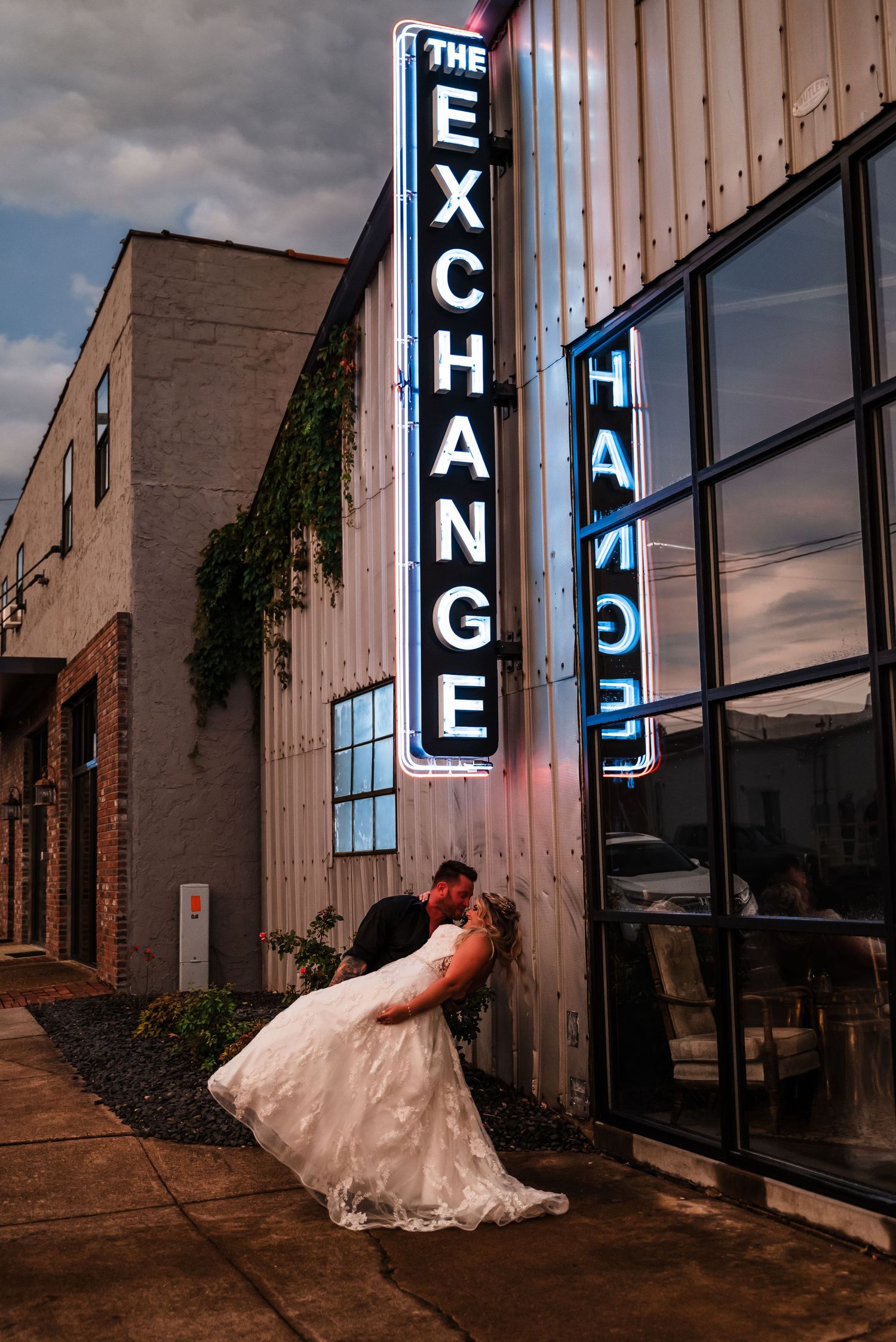 A couple shares a romantic moment outside an urban venue called The Exchange with neon signage at dusk.