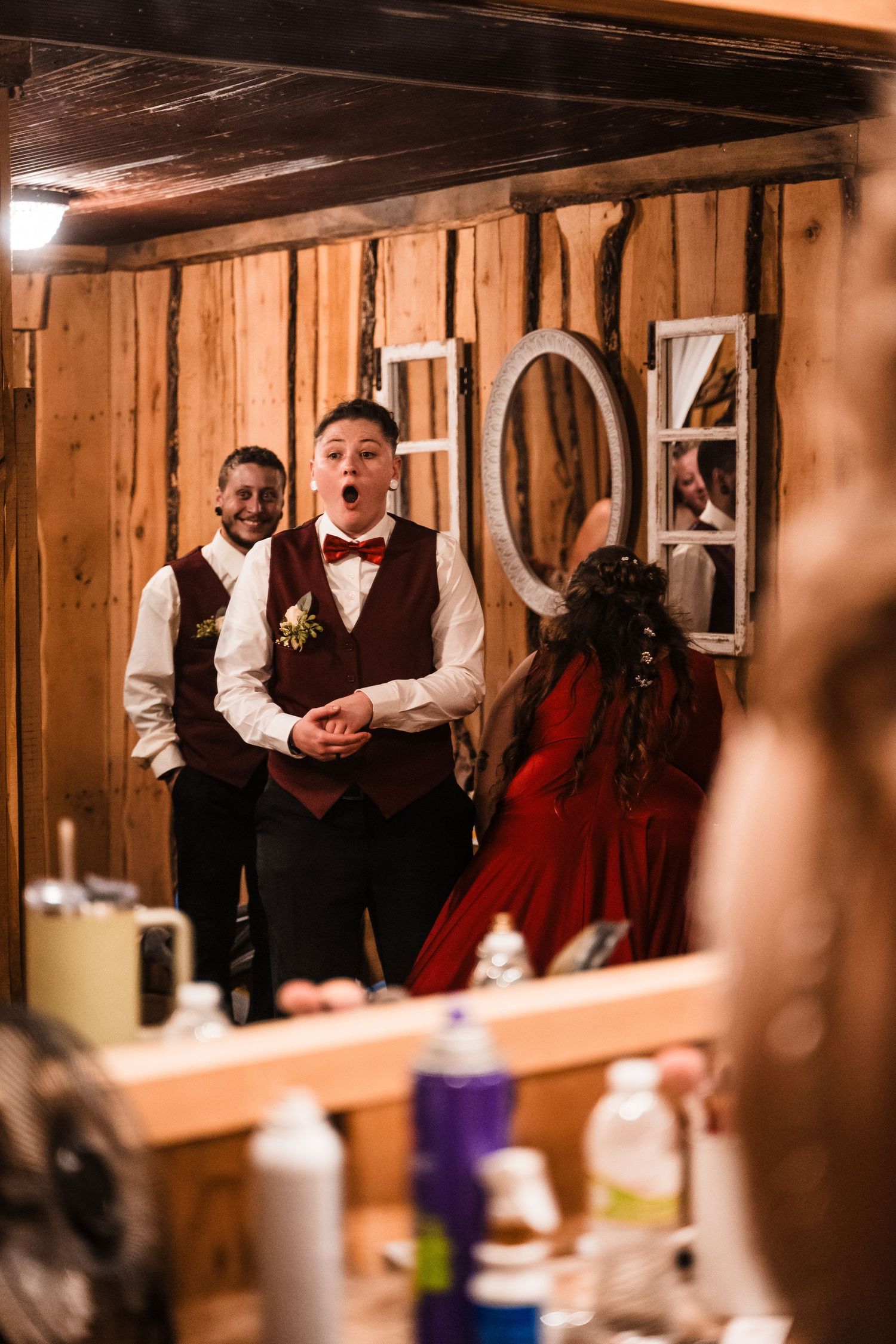 Wedding party members in burgundy vests and bow ties gather in rustic wooden interior.