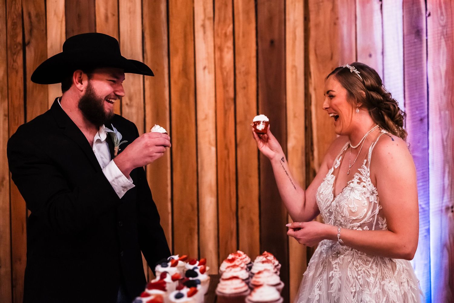 A couple shares a playful moment with cupcakes at a rustic barn wedding reception.
