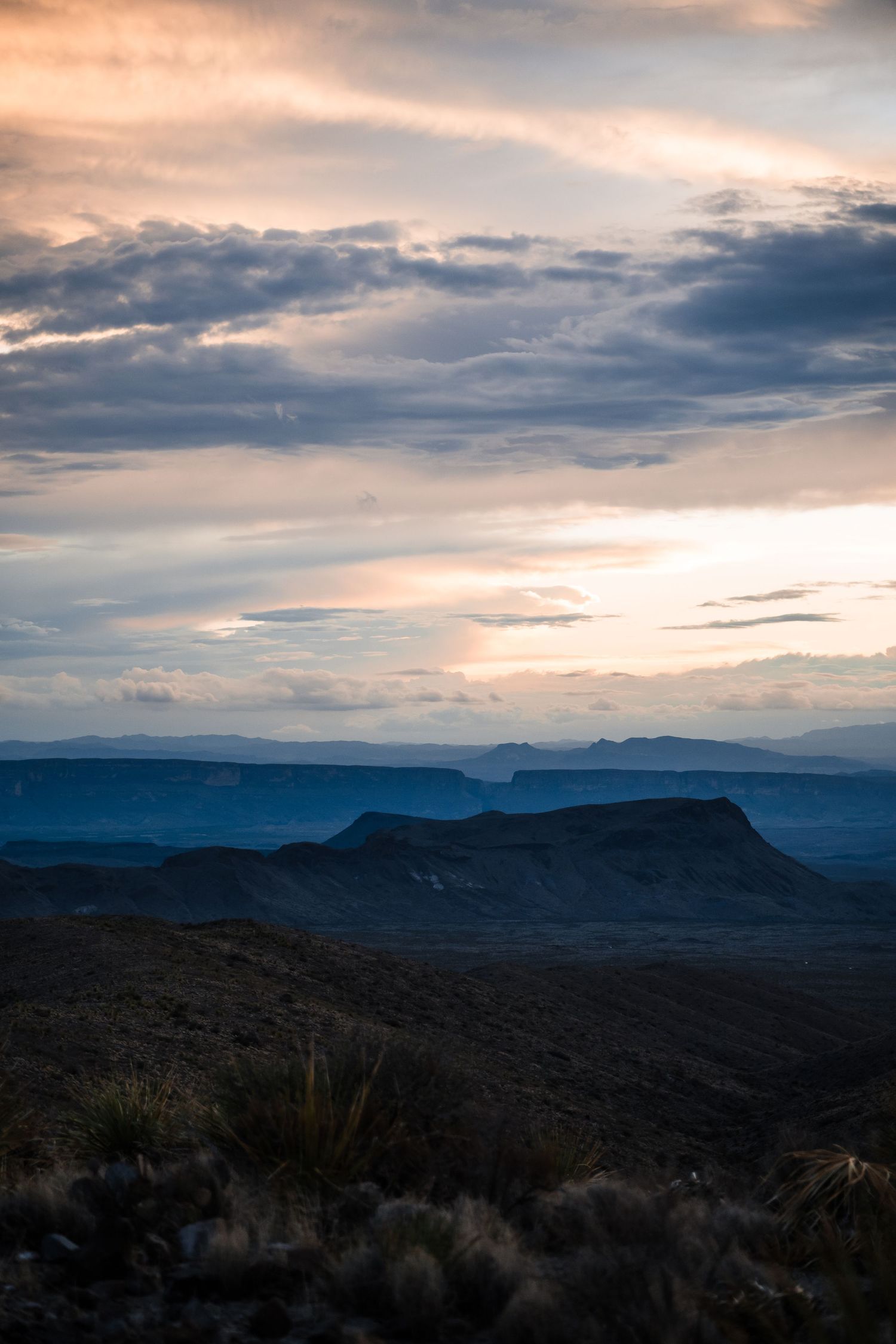 Dramatic sunset over rugged desert mountains with layered blue ridges and moody clouds in the distance.