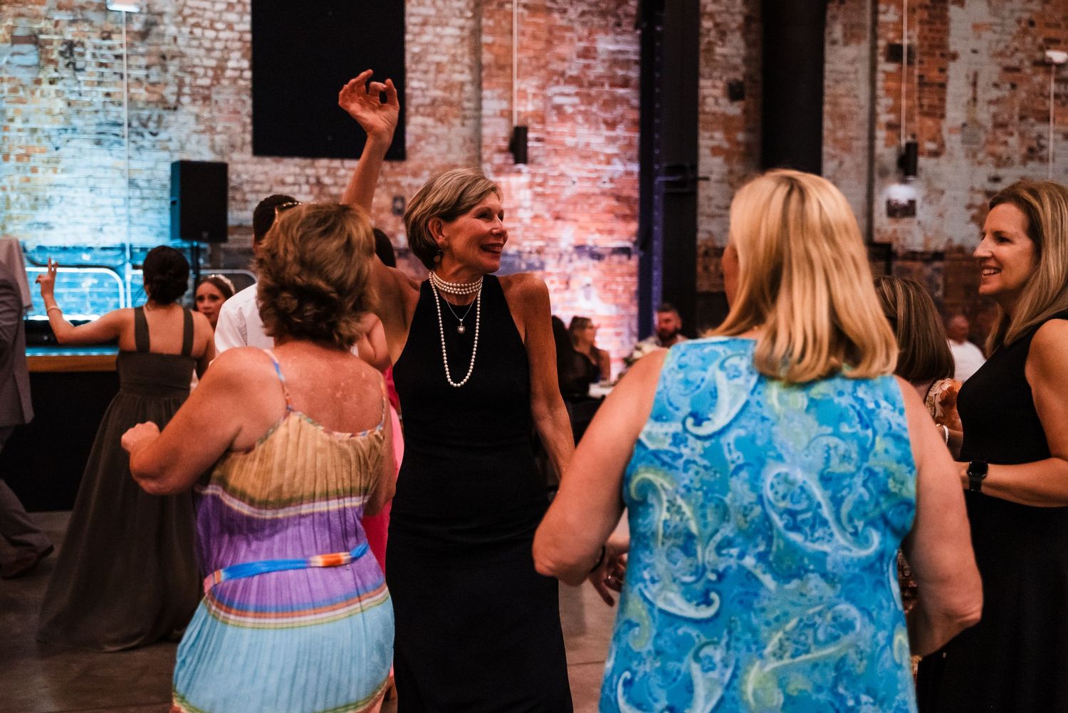 Wedding guests dance and celebrate on the dance floor at an indoor reception with exposed brick walls.