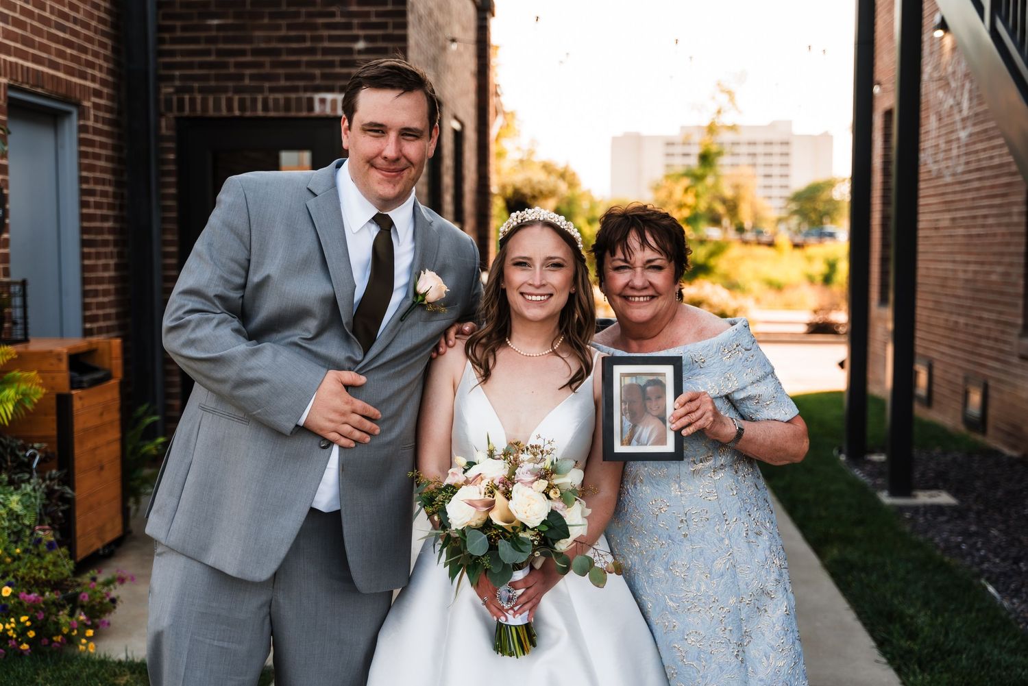 Three people pose together outdoors at a wedding celebration with a bouquet and framed photo near brick buildings.