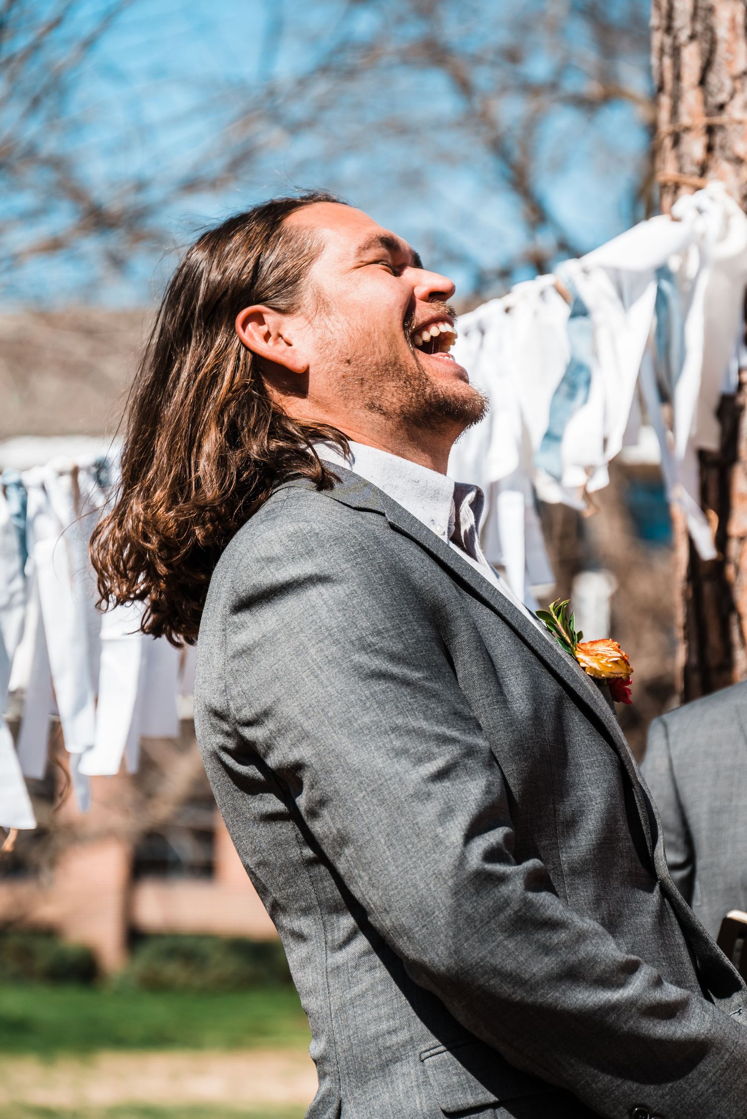 A person in a grey suit jacket laughs joyfully outdoors with white decorations in the background.