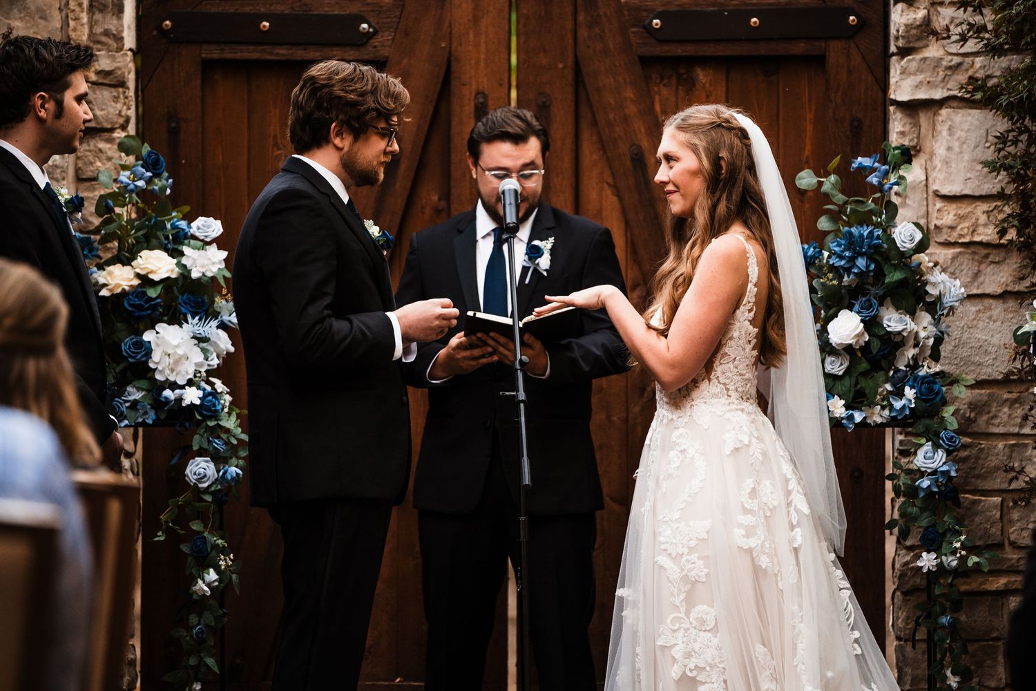 Wedding ceremony takes place in front of rustic wooden doors decorated with blue flowers and string lights.