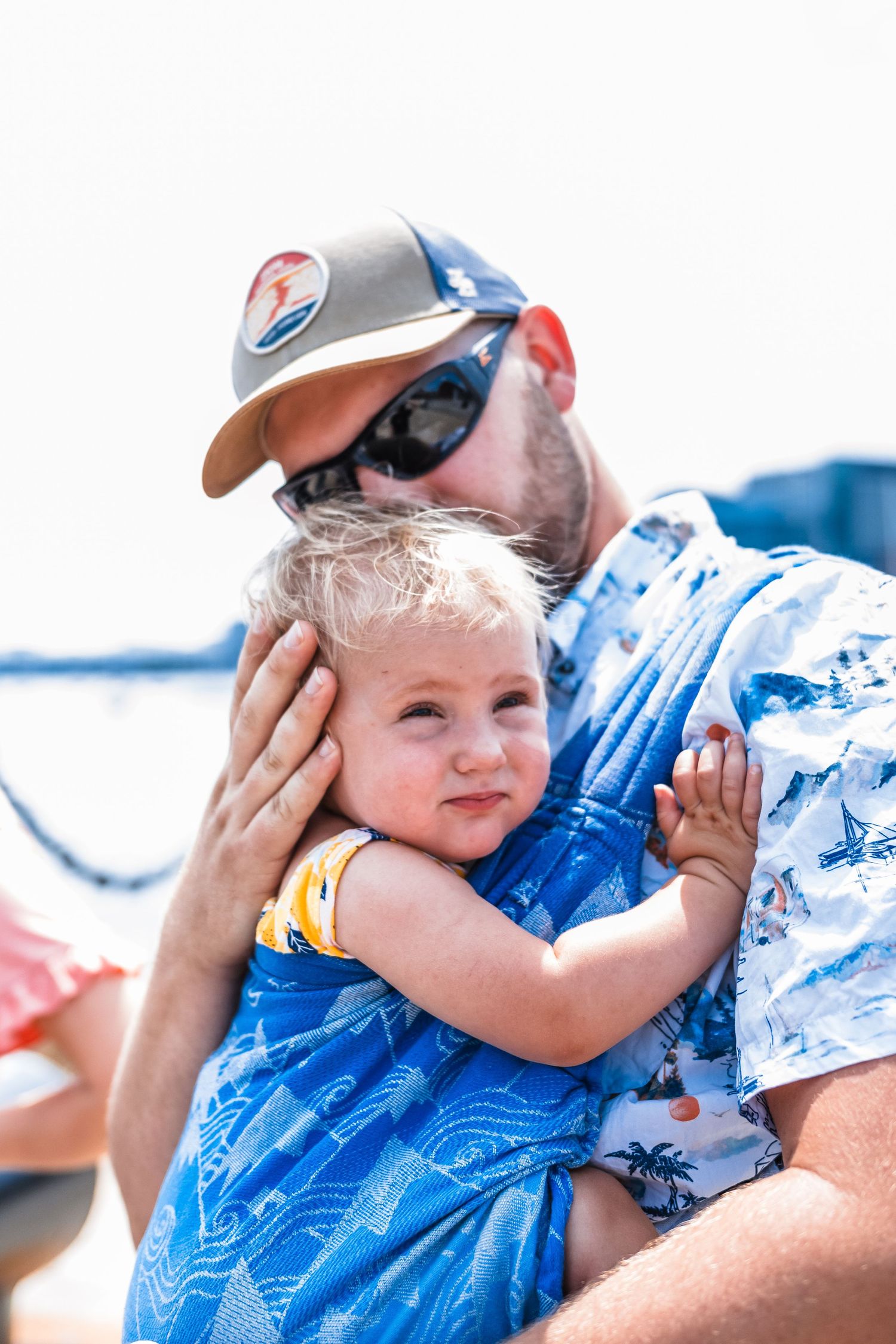 Baby in blue outfit being held close while enjoying sunny day by waterfront.