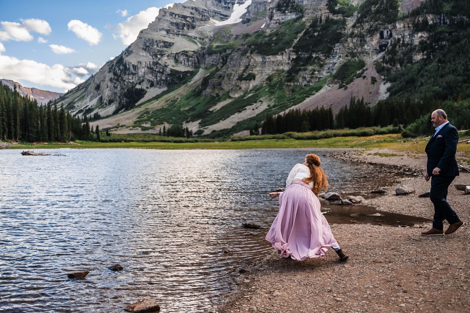Couple walking along rocky mountain lakeshore with majestic peaks in the background.