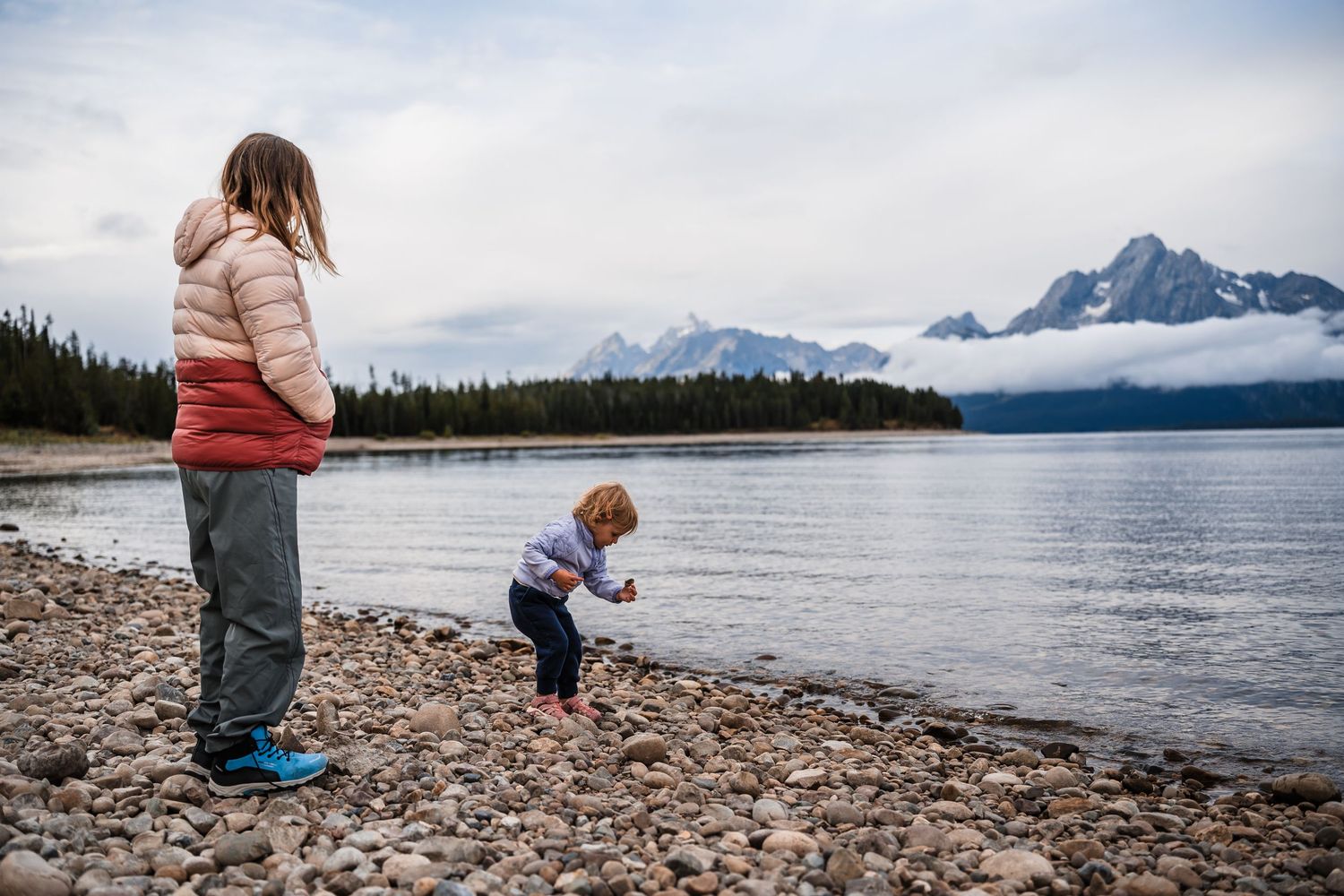 People exploring the rocky shoreline of a mountain lake with snow-capped peaks in the background.