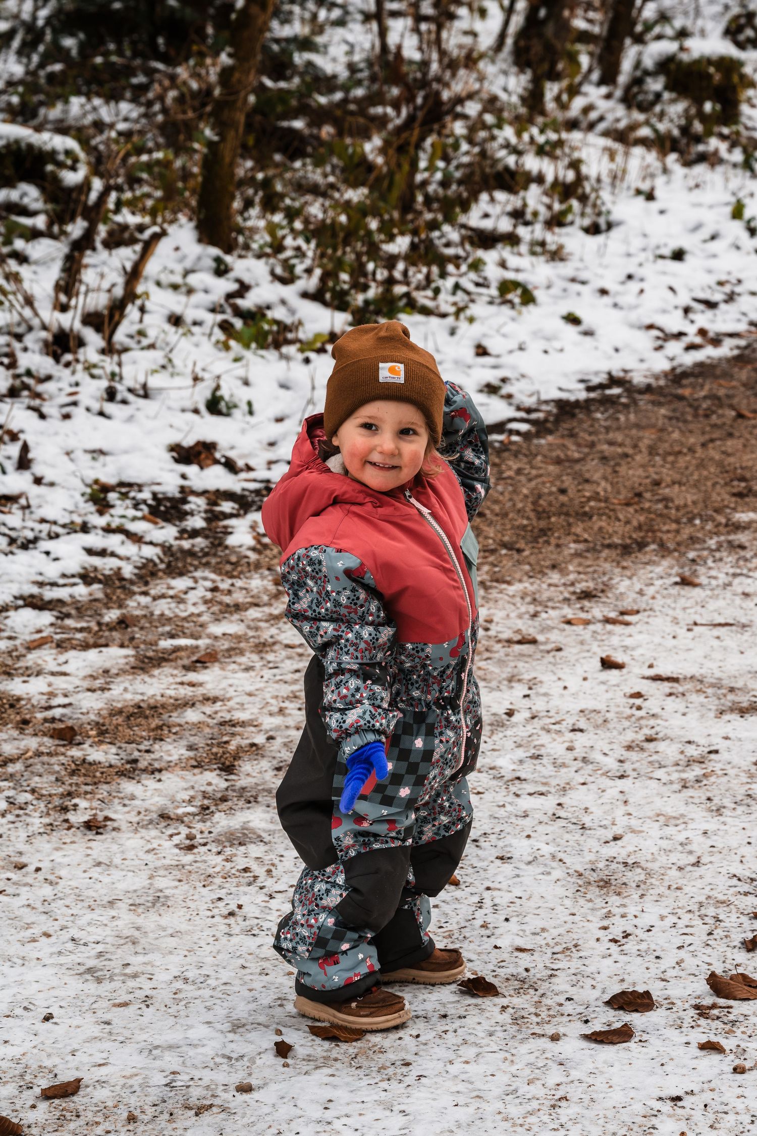Small child wearing winter gear stands on a snowy trail in the woods wearing a brown beanie and patterned snowsuit.