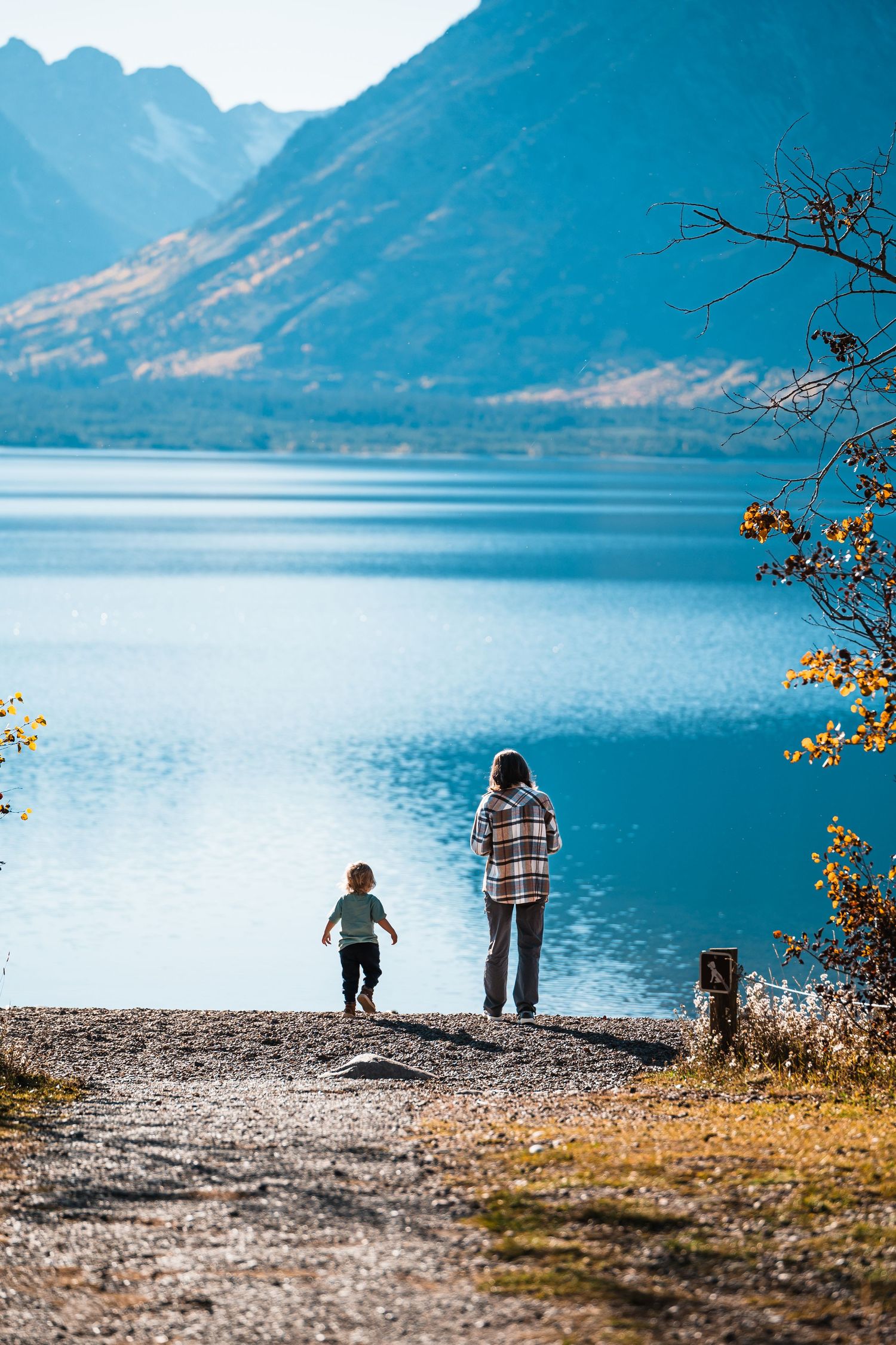 Silhouettes of people standing on a rocky shoreline against turquoise lake waters and mountain backdrop.