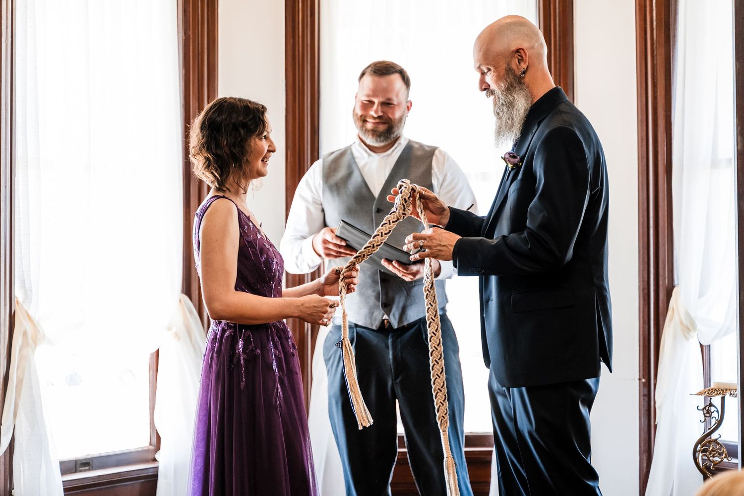 Handfasting ceremony with officiant binding couple's hands with rope indoors near bright windows.