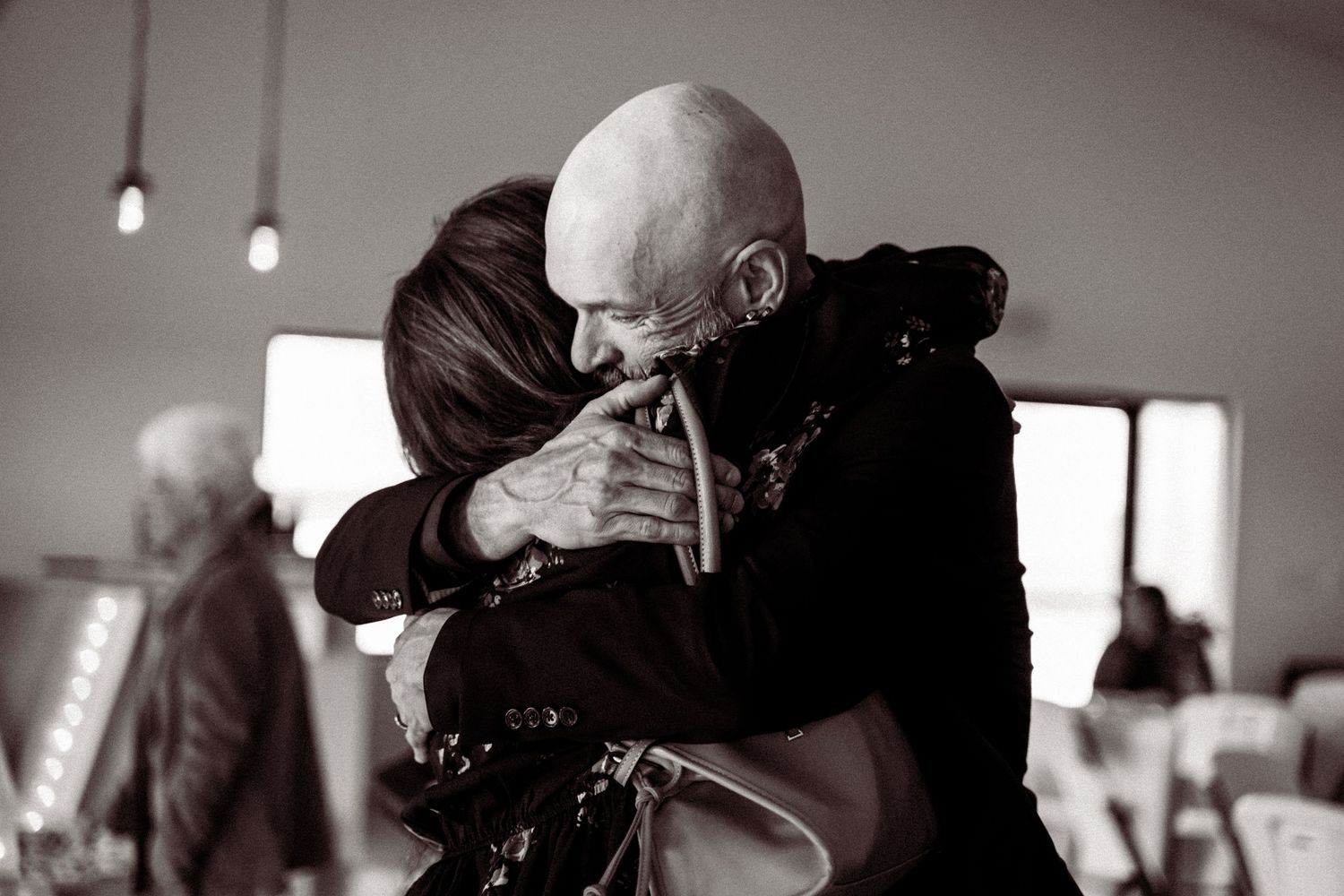Black and white photo of a bald man and woman sharing a heartfelt embrace indoors.