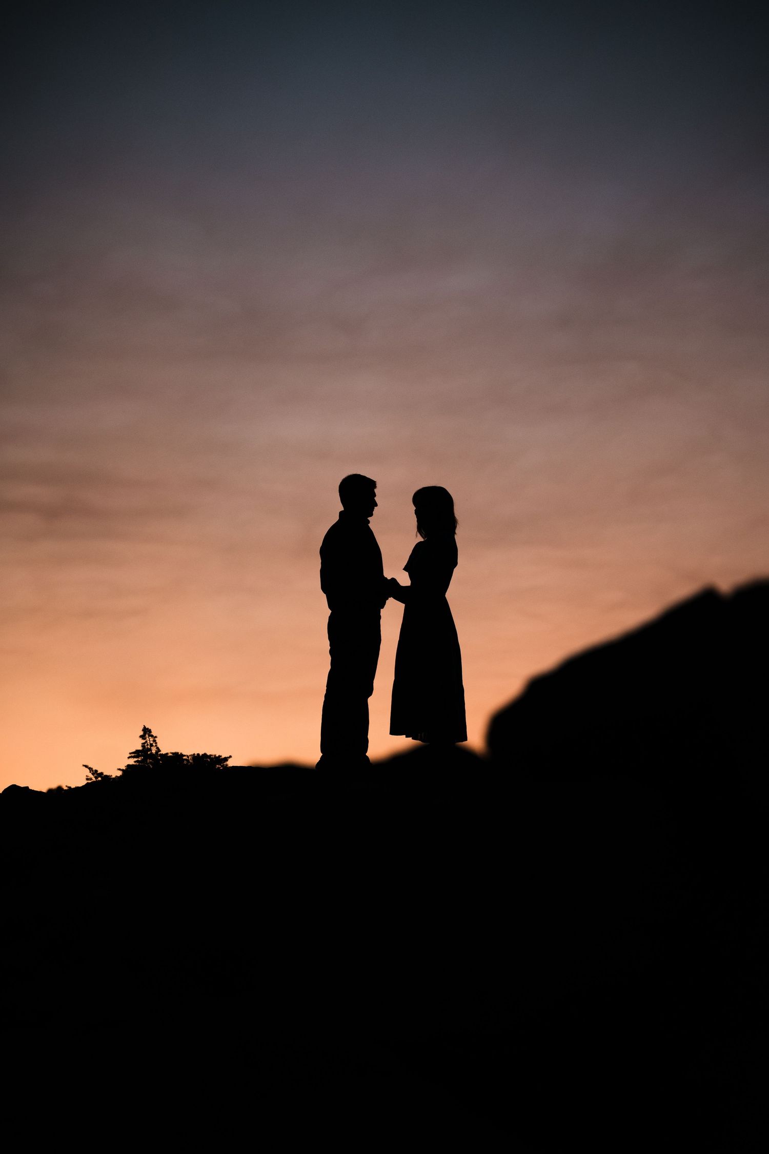Silhouettes of two figures stand together against a dramatic sunset sky creating a romantic atmospheric scene.