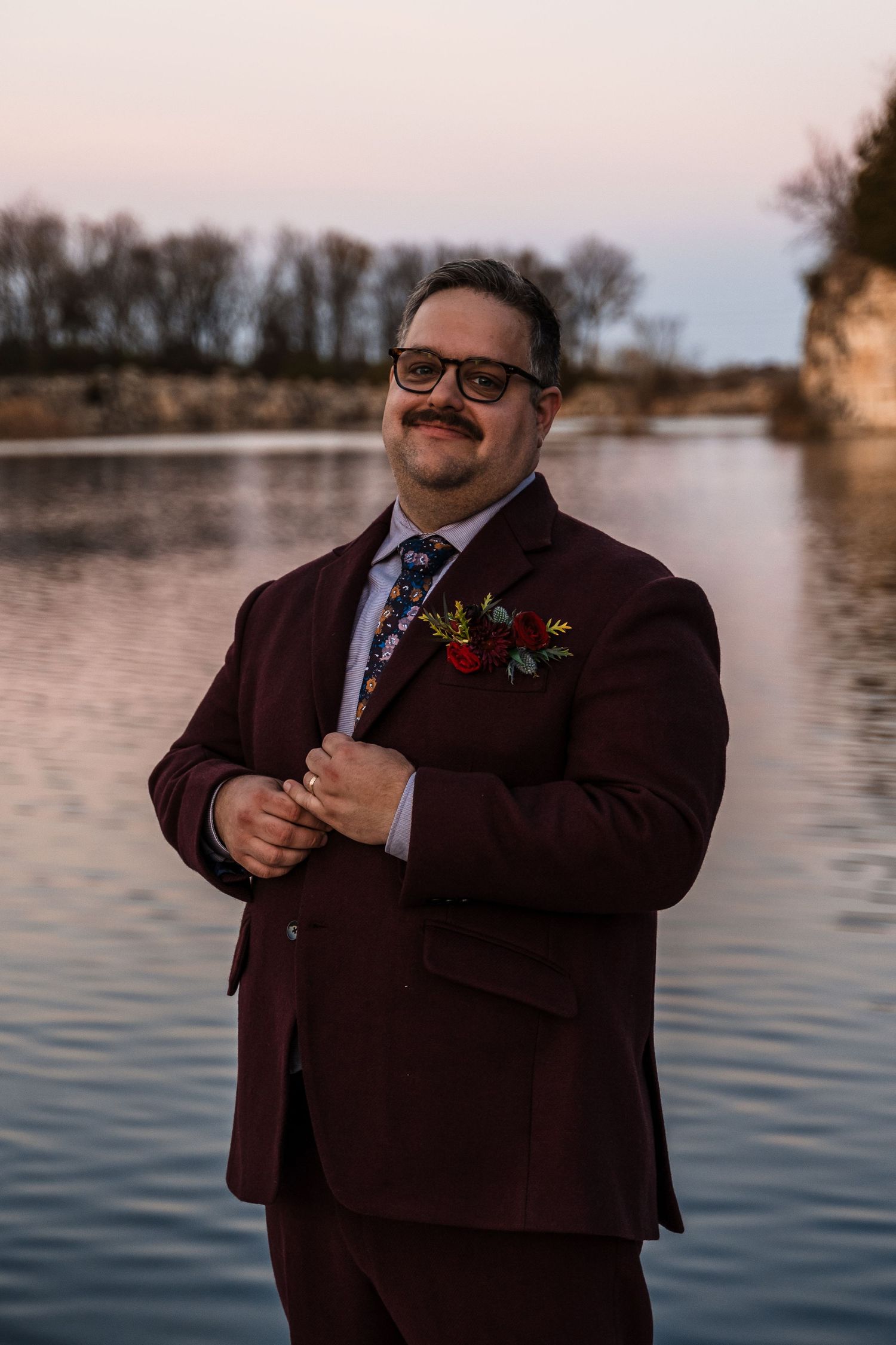 Person in burgundy suit with boutonniere posing playfully by a waterfront at sunset.