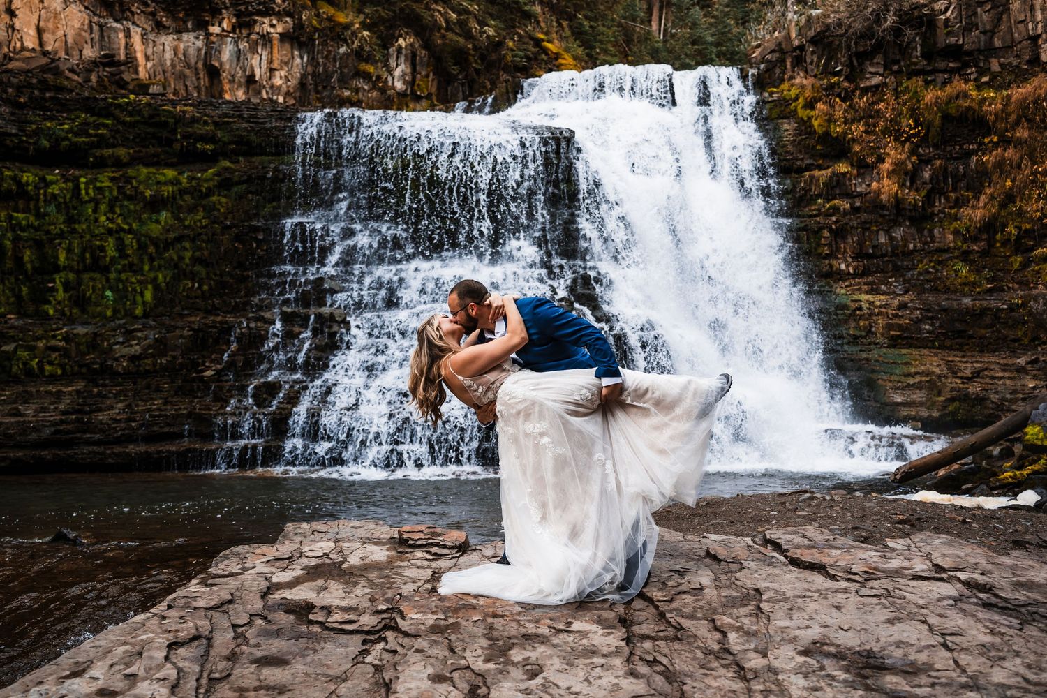 A couple poses for romantic photos in front of a cascading waterfall surrounded by autumn foliage.