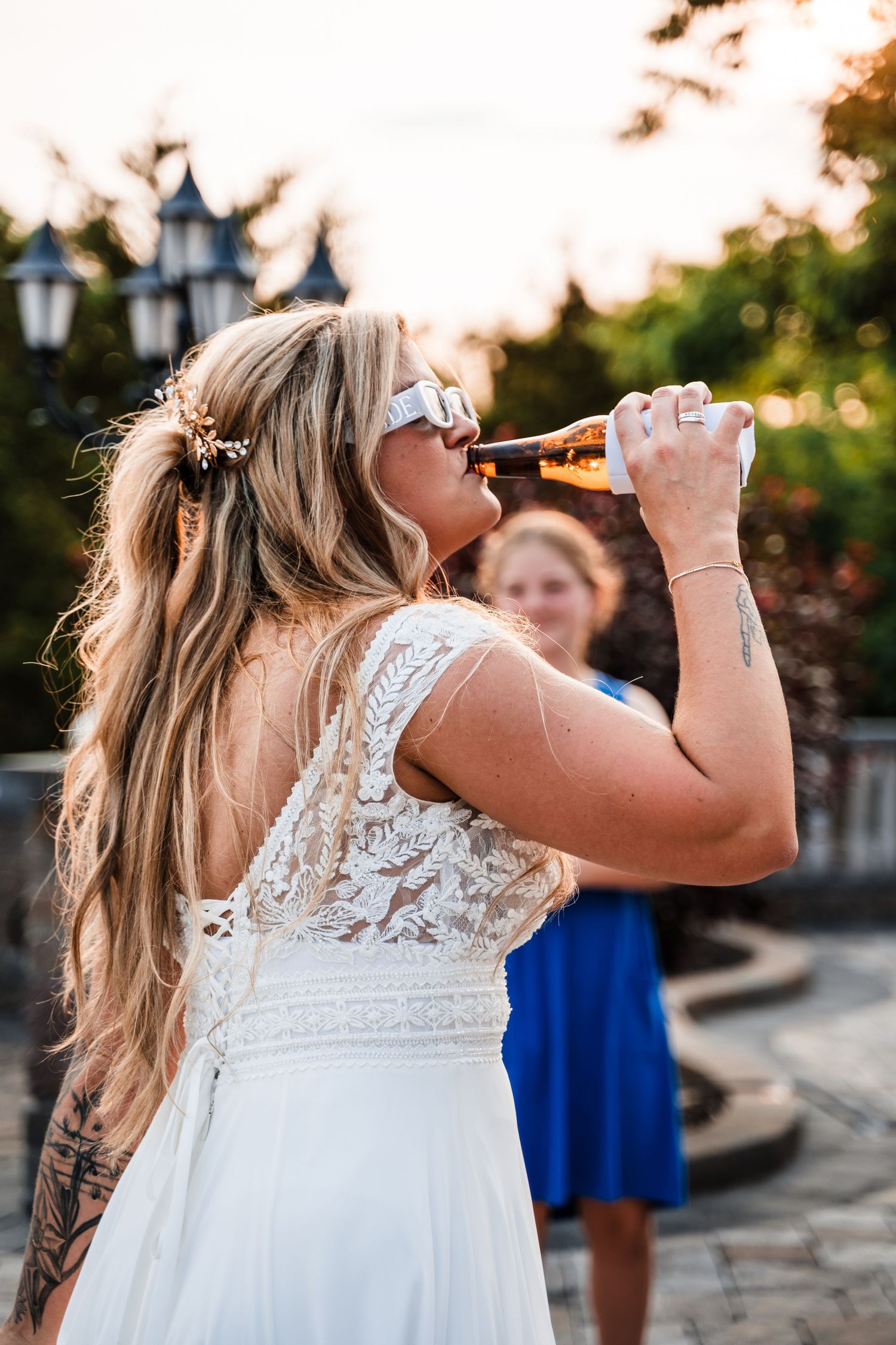 Person in white lace dress takes a drink from a bottle during an outdoor celebration.