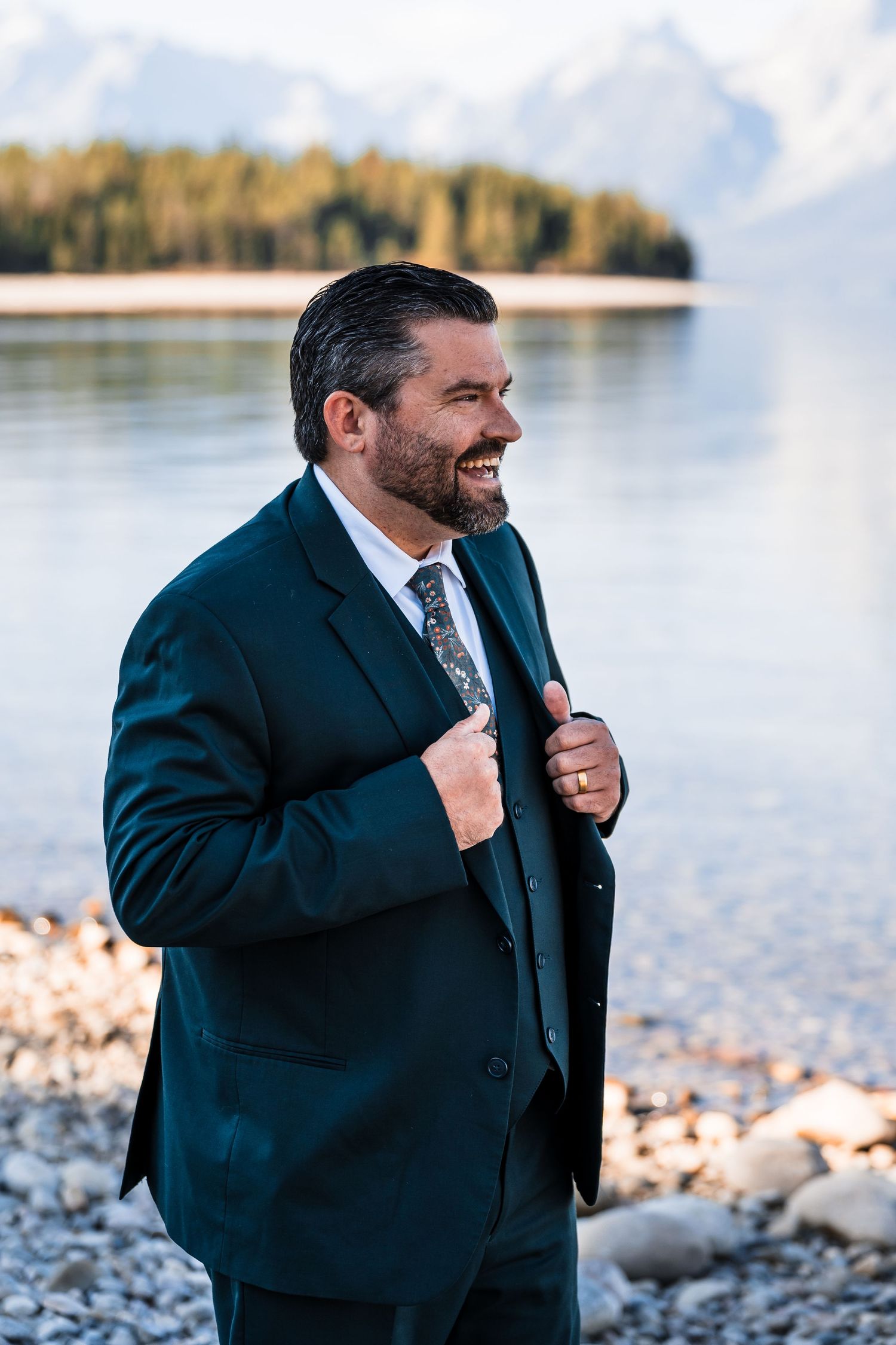 A groom in a dark suit laughs joyfully by a scenic lake during a wedding photoshoot.