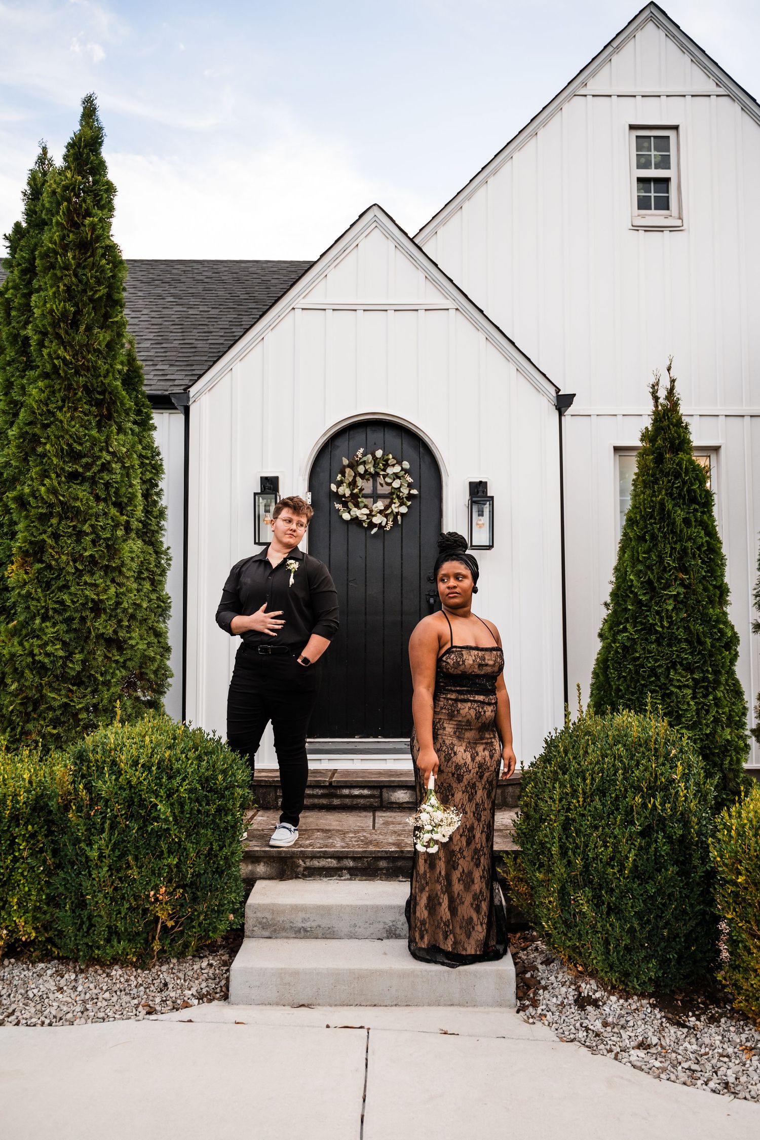 A couple standing in front of a modern white chapel with black door and cypress trees.