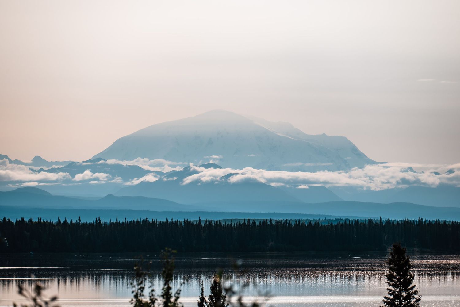 Misty mountain peak rises above a tranquil lake surrounded by evergreen forest silhouettes at dusk.