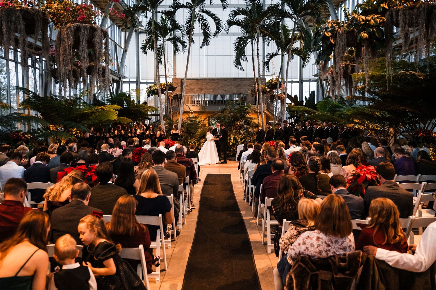 Large indoor wedding ceremony with palm trees and a long aisle lined with white chairs.