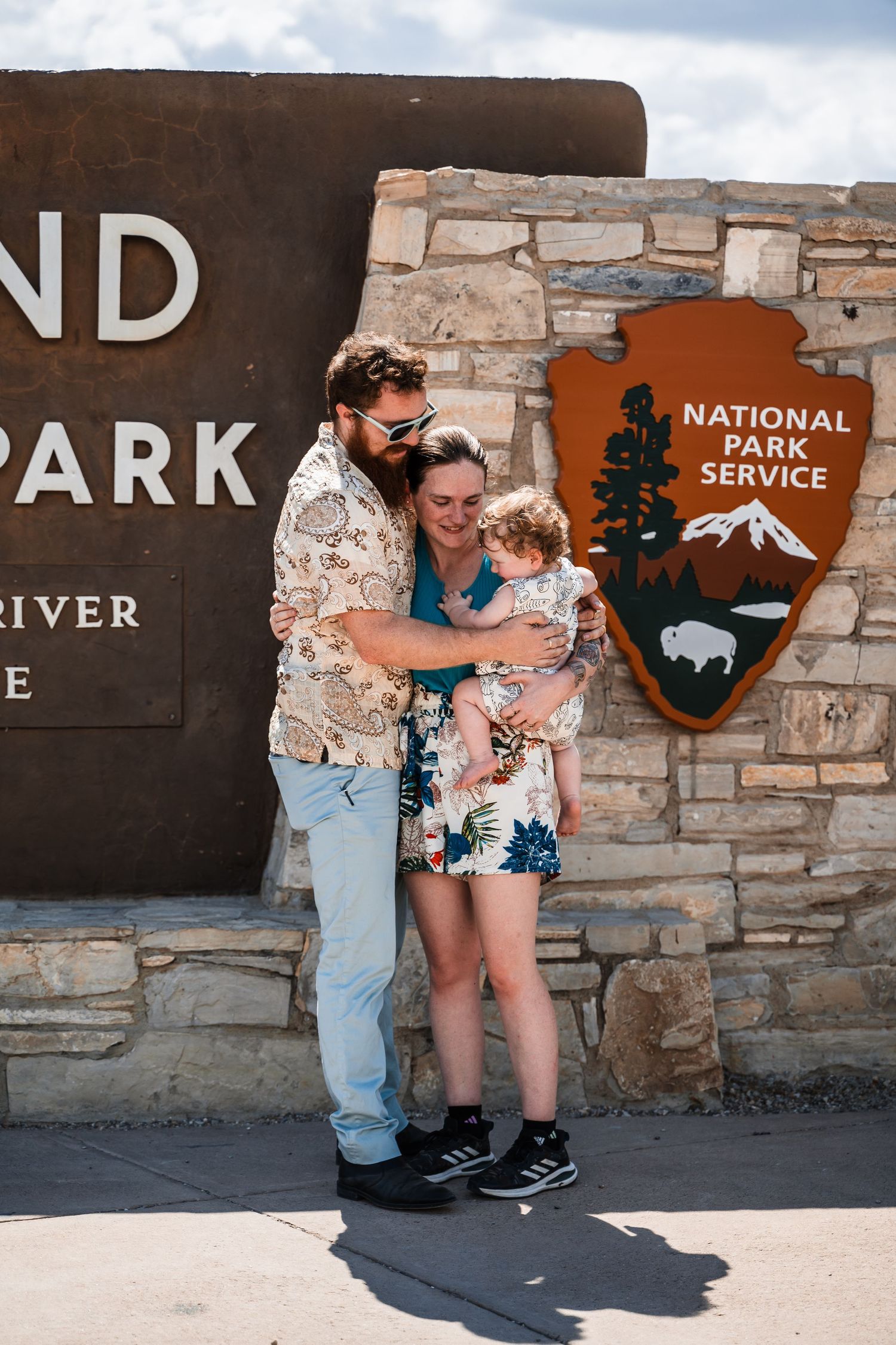 A family poses together in front of a National Park Service sign and stone wall entrance.