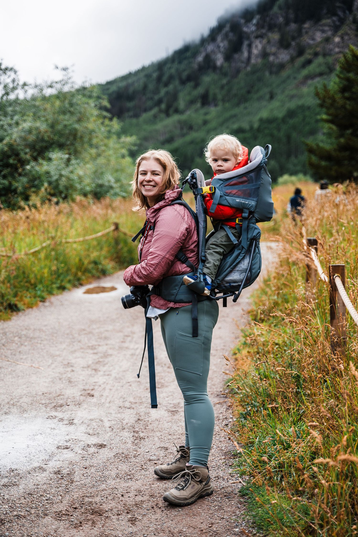 Hiker carrying a child in a backpack carrier on a mountain trail surrounded by lush greenery.