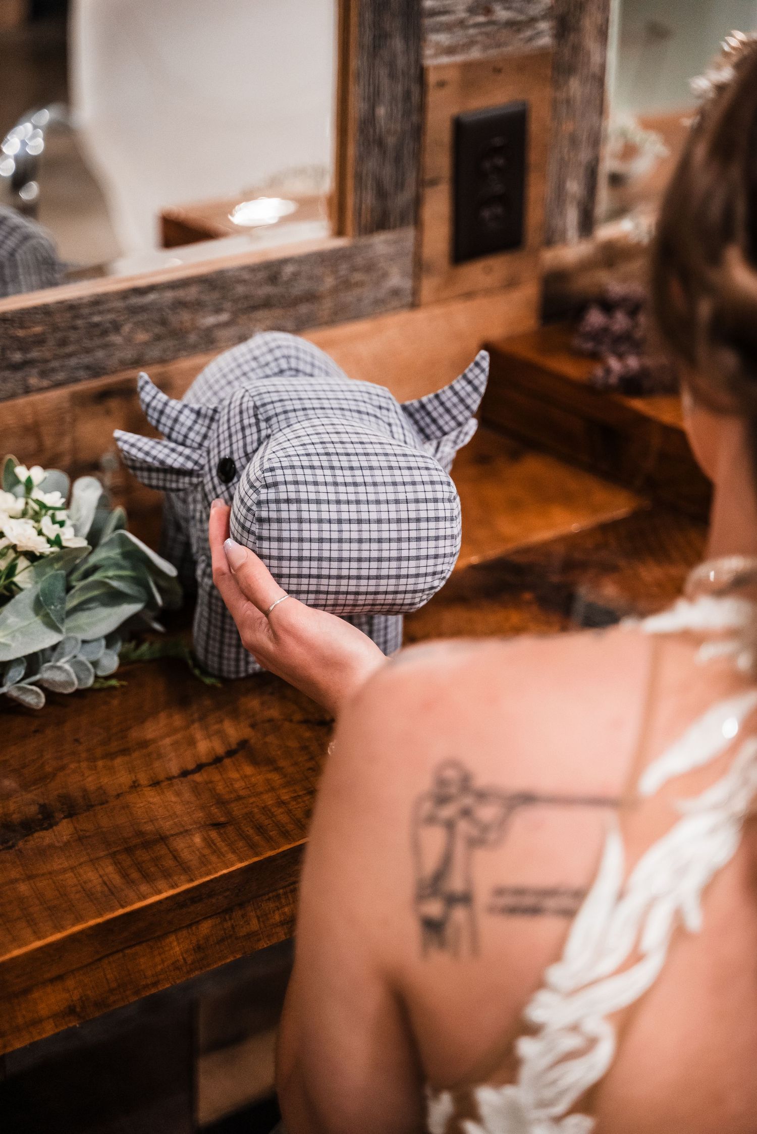 A tattooed arm holds a plaid stuffed toy with horns next to a wooden vanity with greenery.