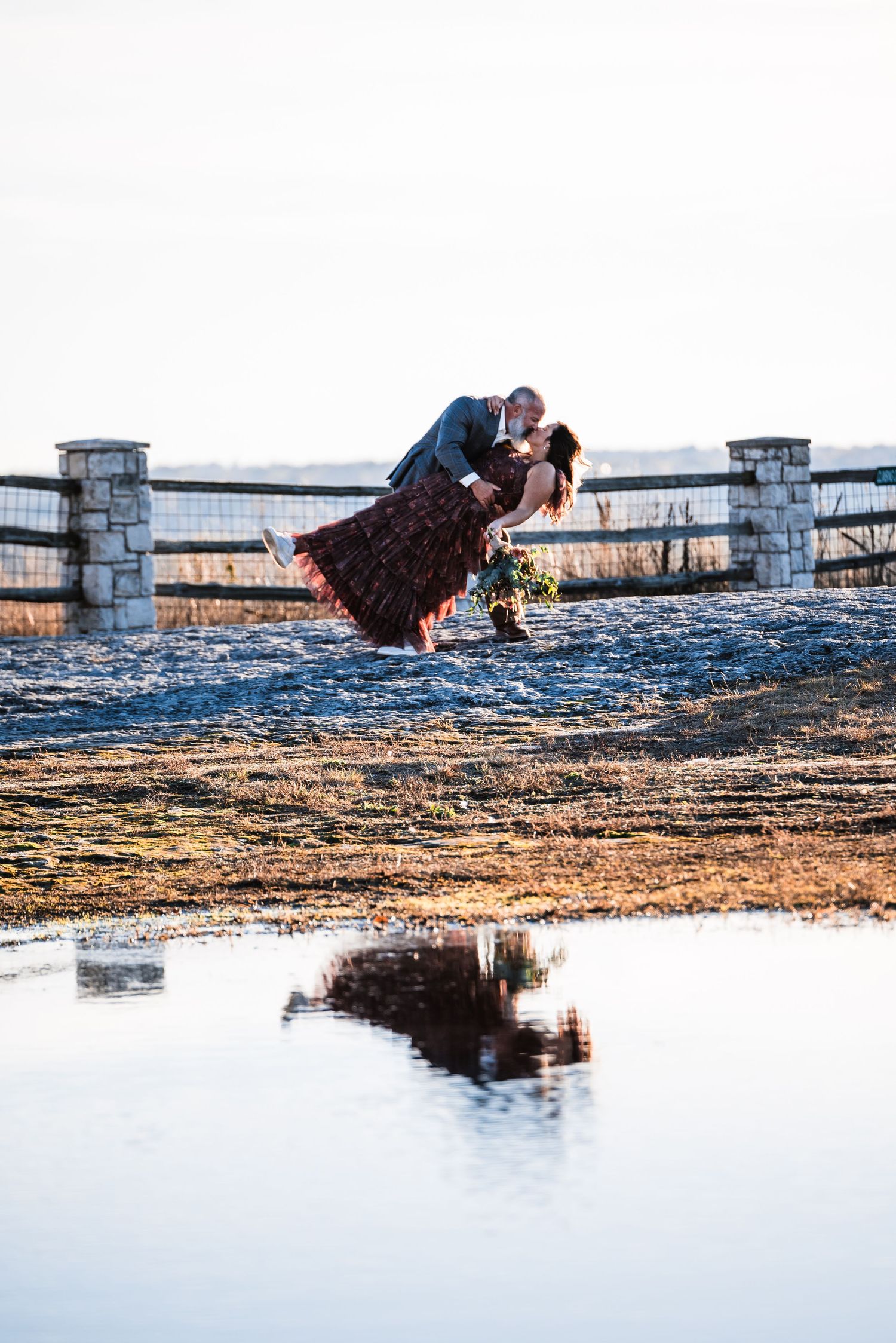 Romantic dip pose by stone wall with water reflection during golden hour.