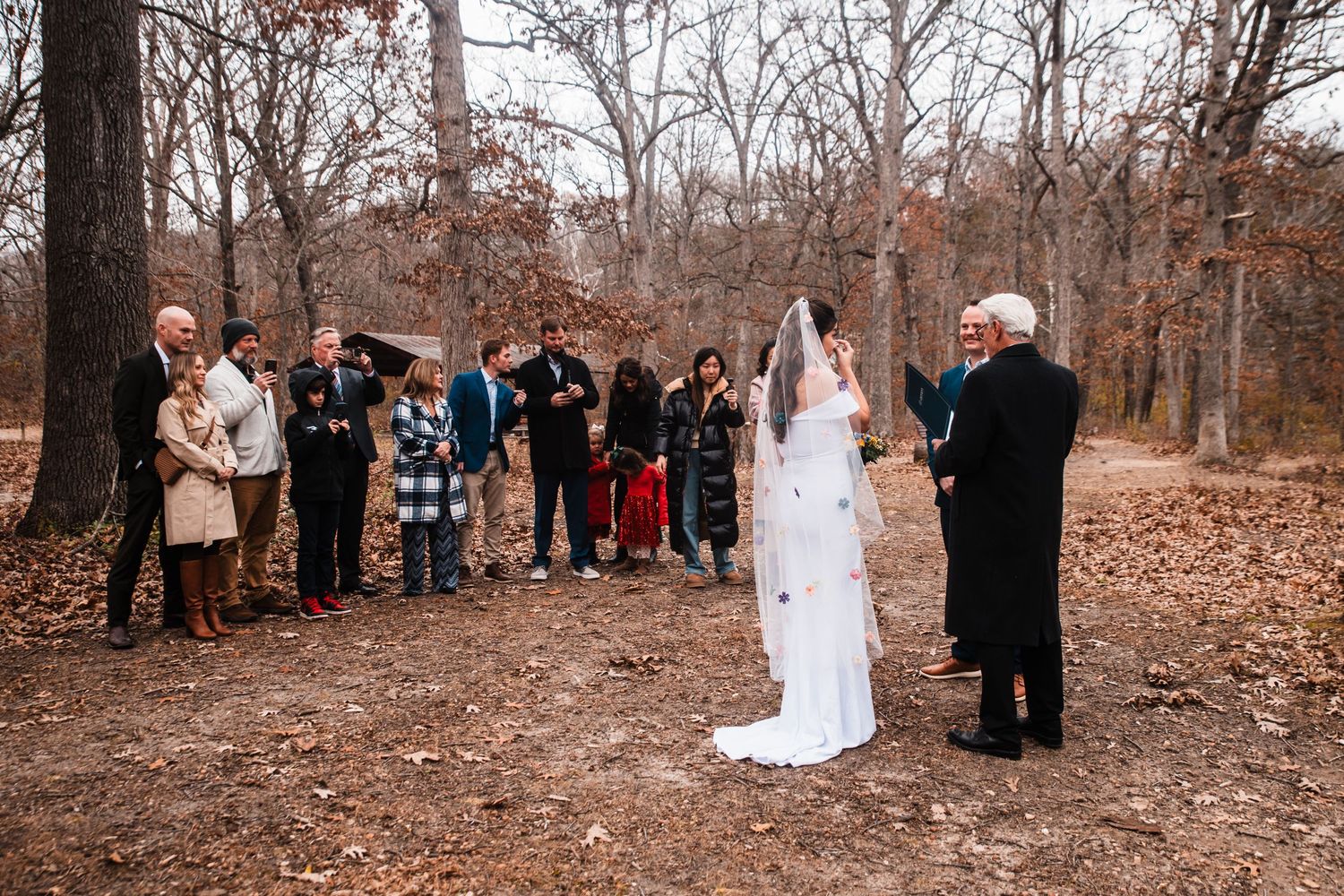 Small wedding ceremony gathered in a wooded area with fall foliage and fallen leaves on the ground.