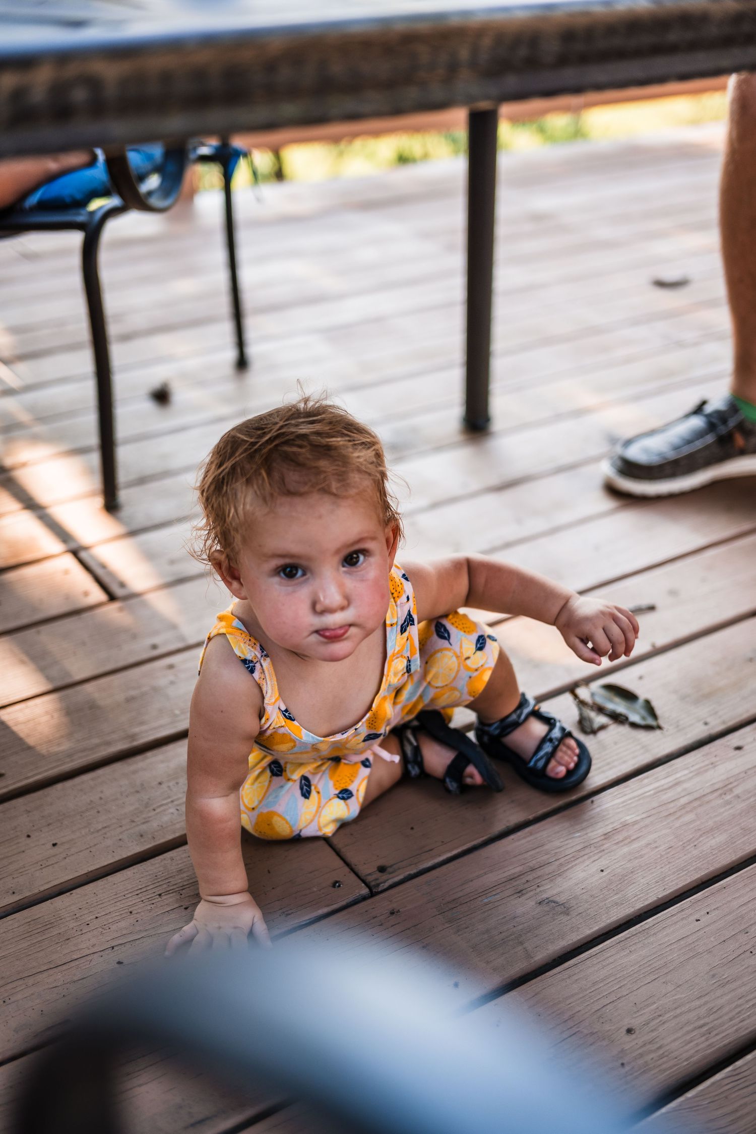 Small child in yellow floral dress sitting on wooden deck in late afternoon sunlight.