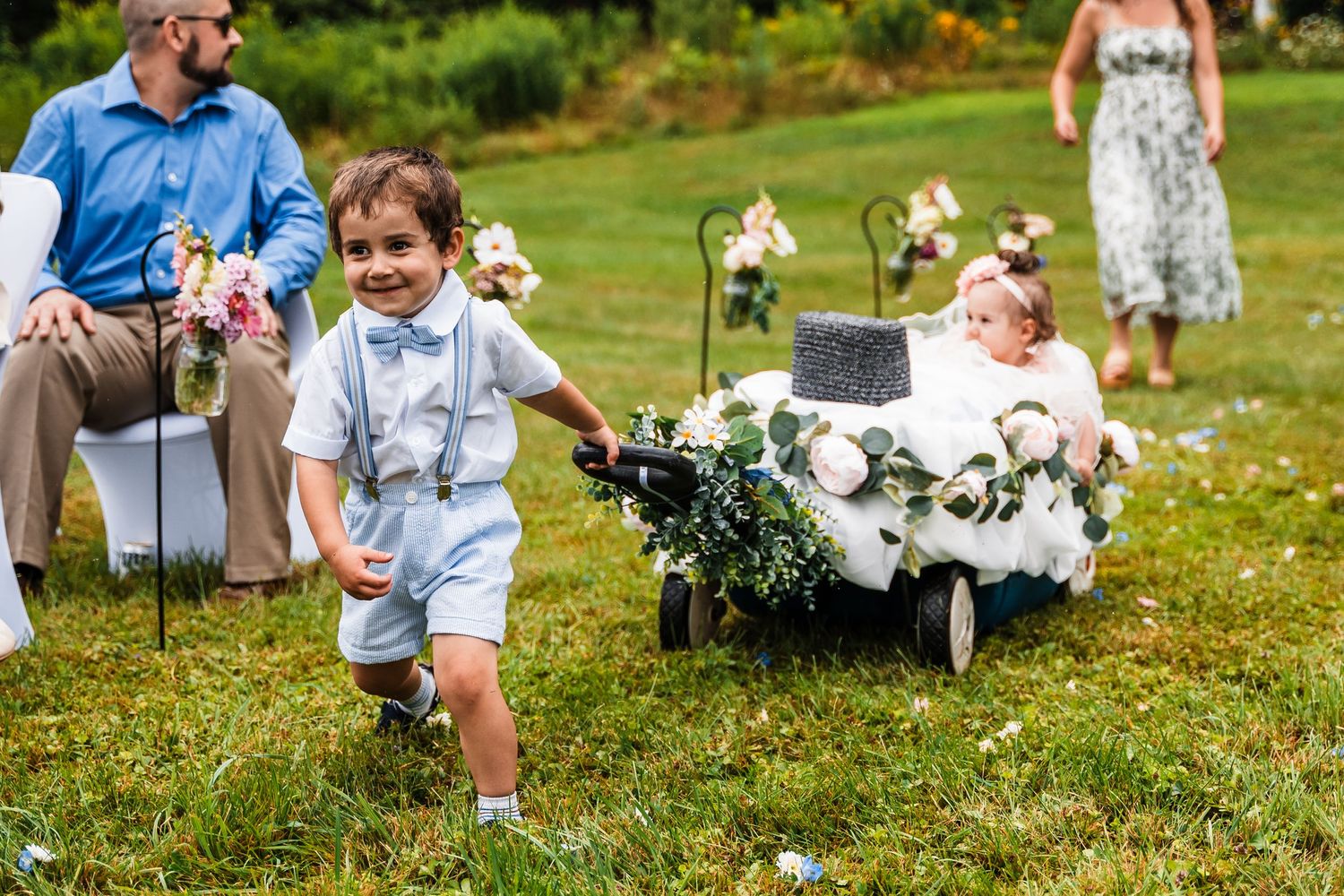 Young ring bearer in blue outfit runs across grass while pulling decorated wagon.