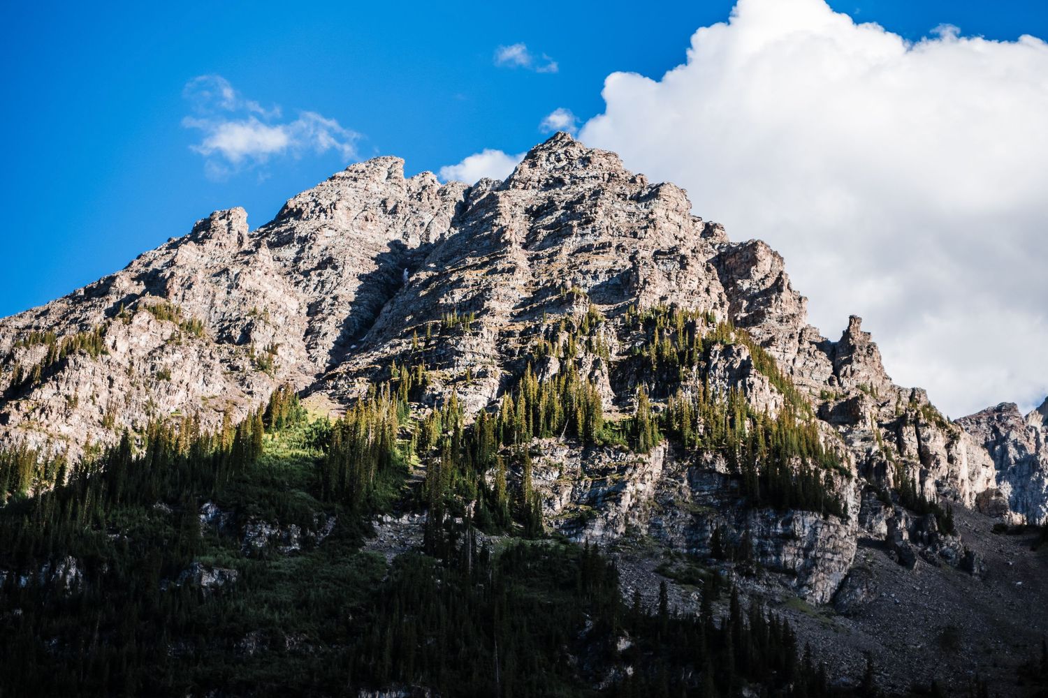 Dramatic rocky mountain peak towers over forest landscape against blue sky and clouds.