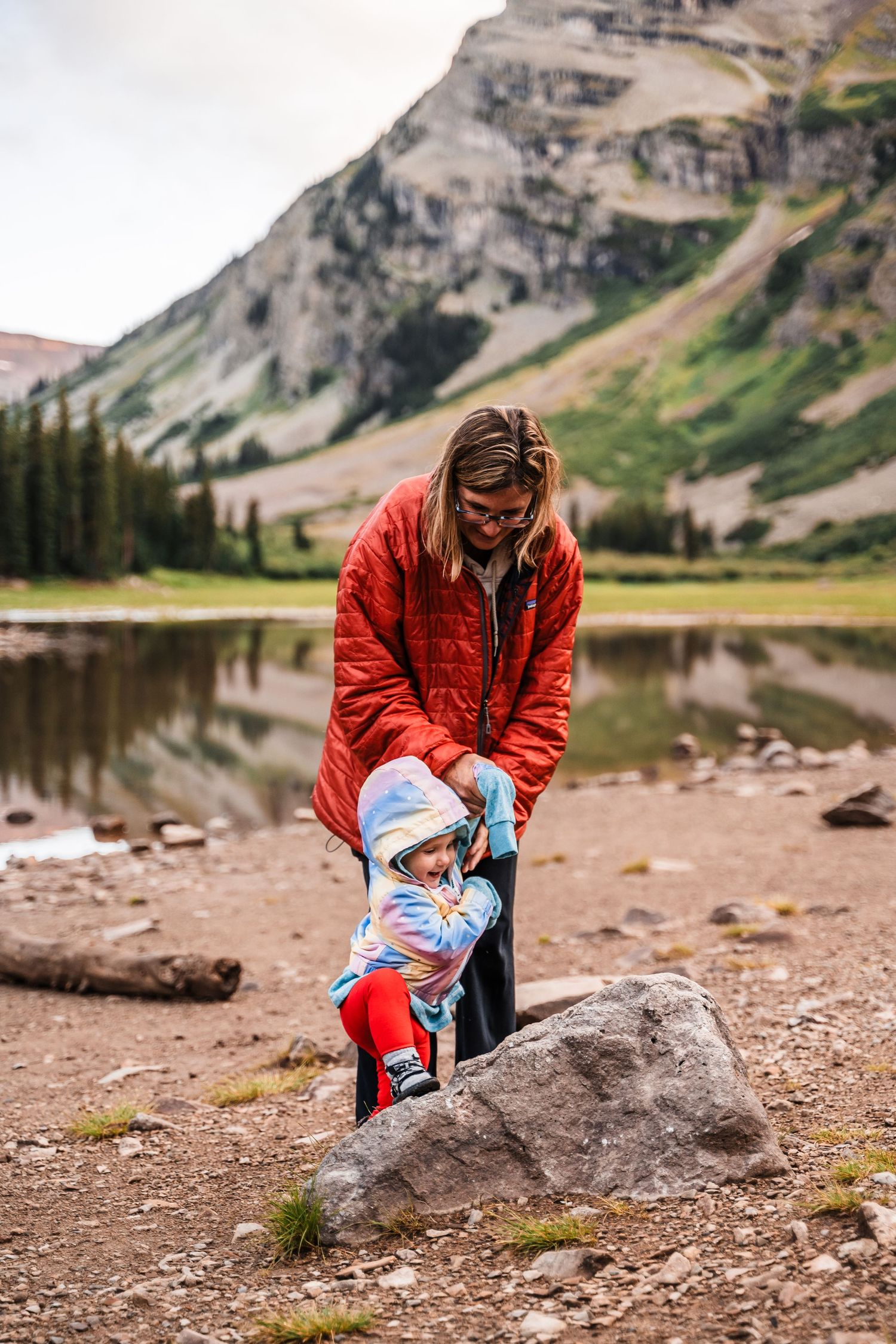 Adult helping child climb rock by mountain lake with dramatic peaks in background.