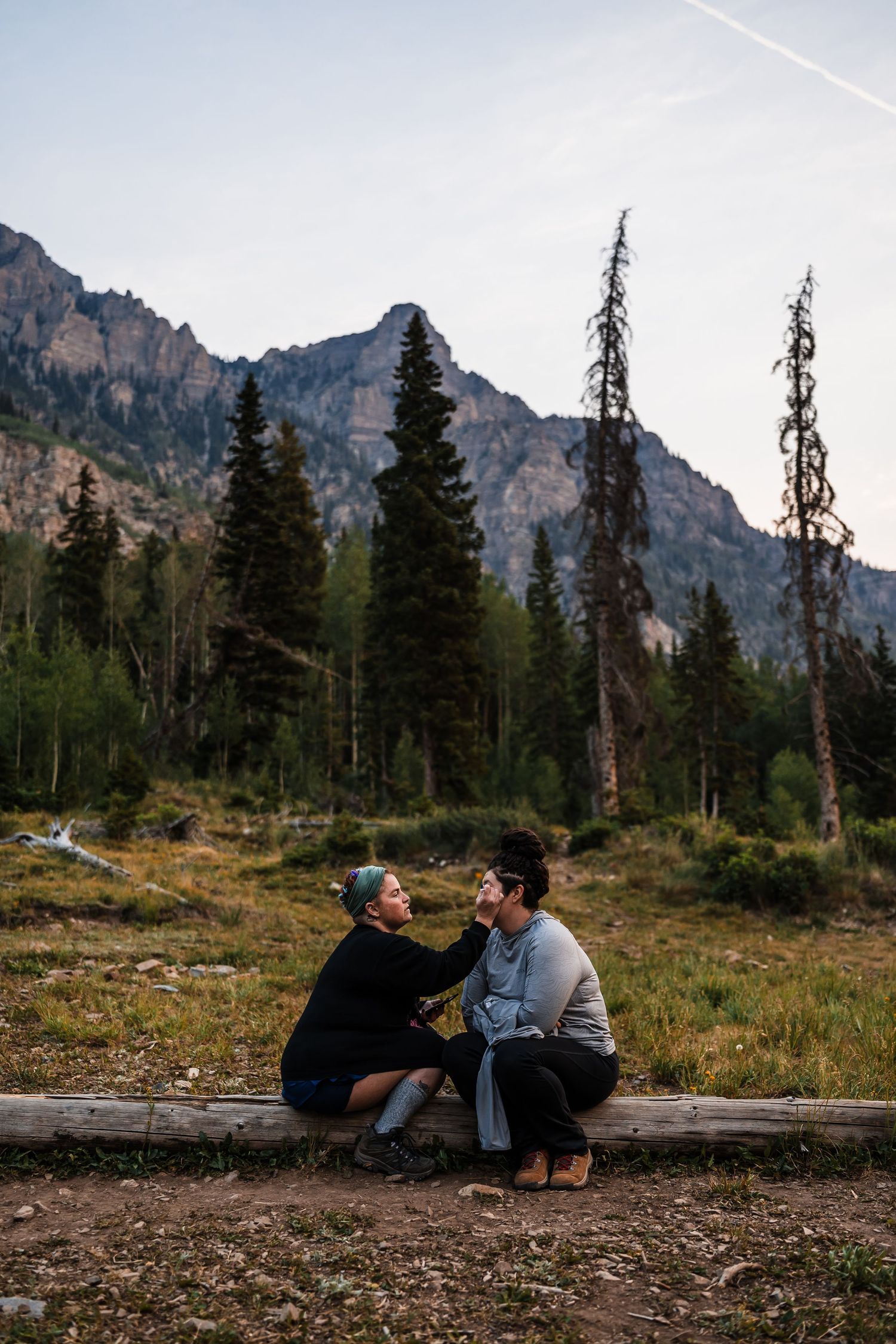 Couple shares tender moment sitting on log with rocky mountain peaks and pine forest backdrop.