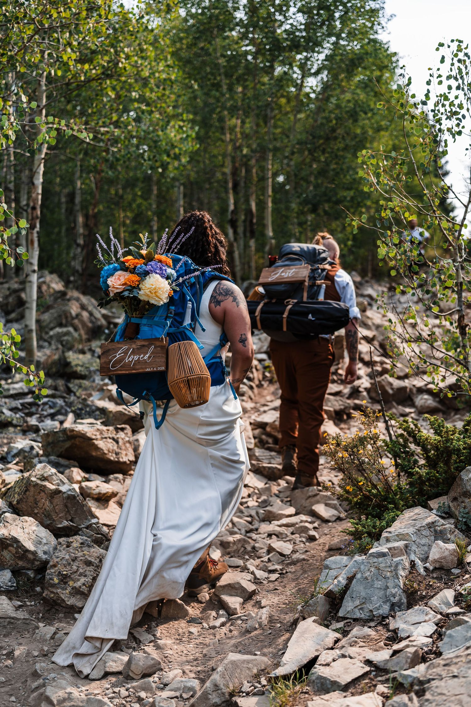 Bride in white gown with floral backpack hikes rocky mountain trail with groom.