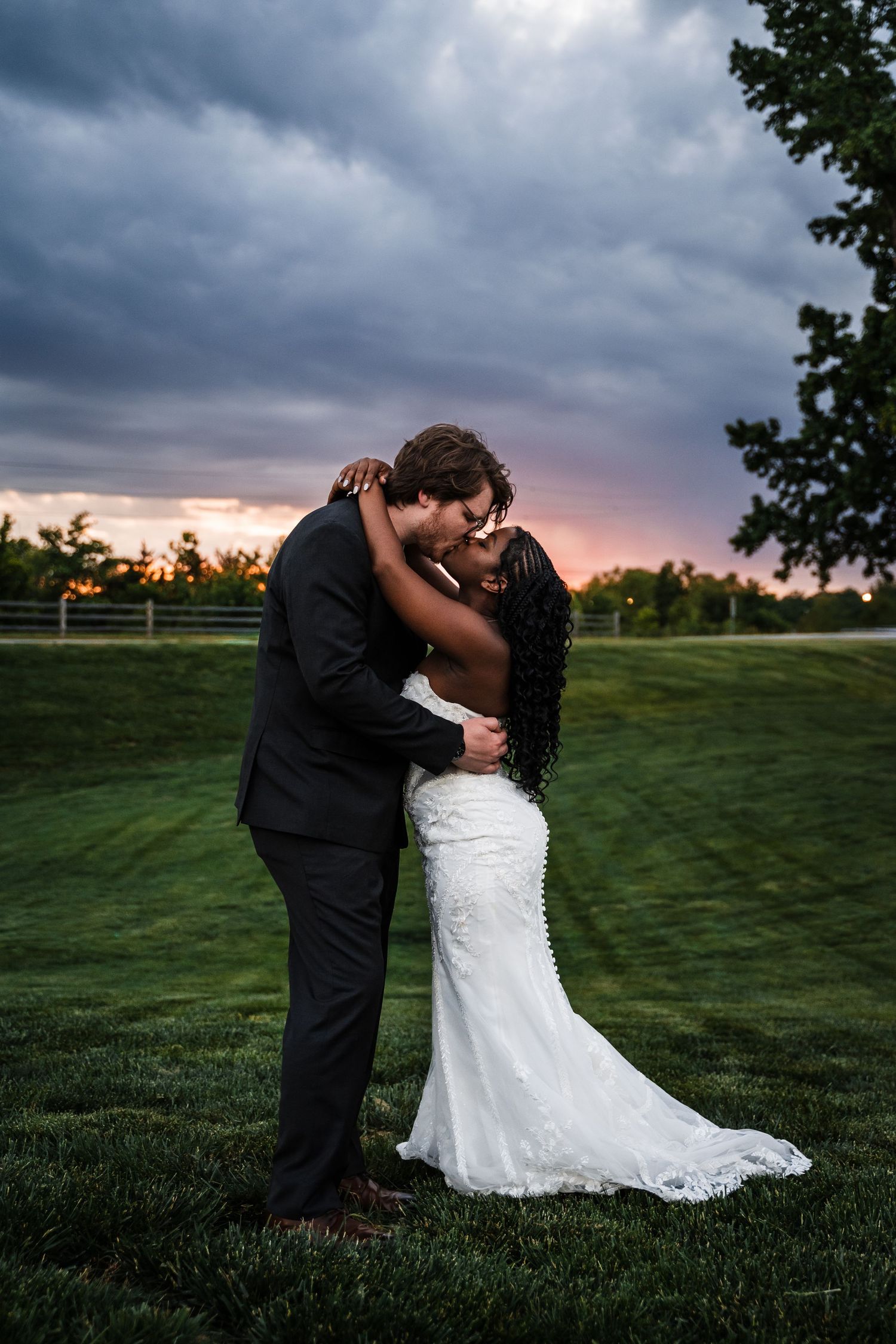 Interracial couple shares romantic kiss in lace wedding gown and dark suit against dramatic sunset sky.