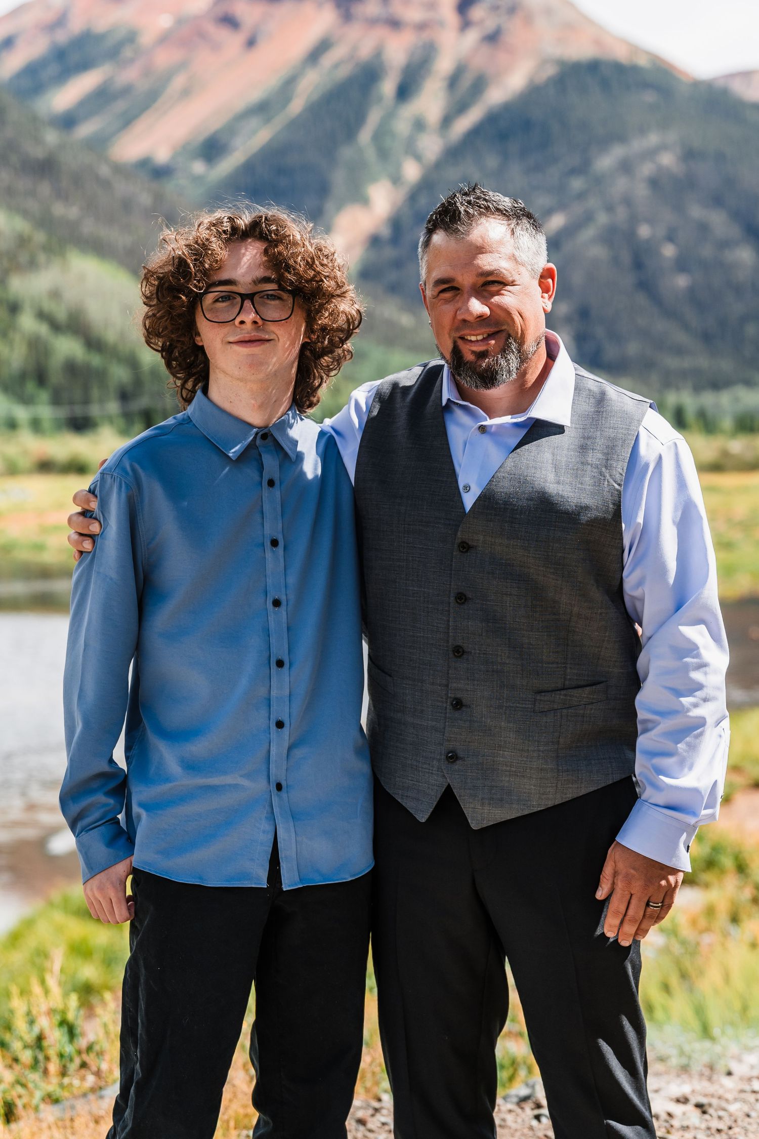 Teenage boy and older man in wedding vest smiling together with mountain and lake backdrop.