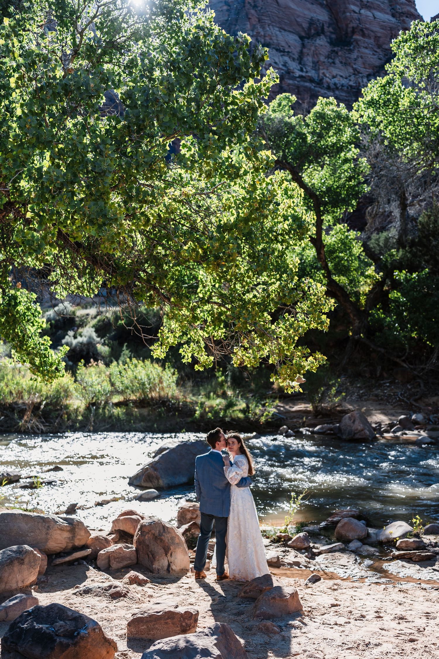 Wedding couple embraces by a rocky river beneath lush green trees in a scenic canyon landscape.
