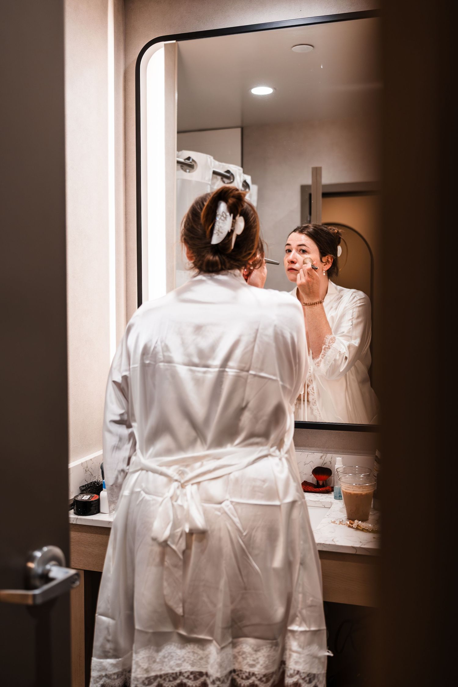 Bride in white satin robe applying makeup at vanity mirror while getting ready for wedding day.