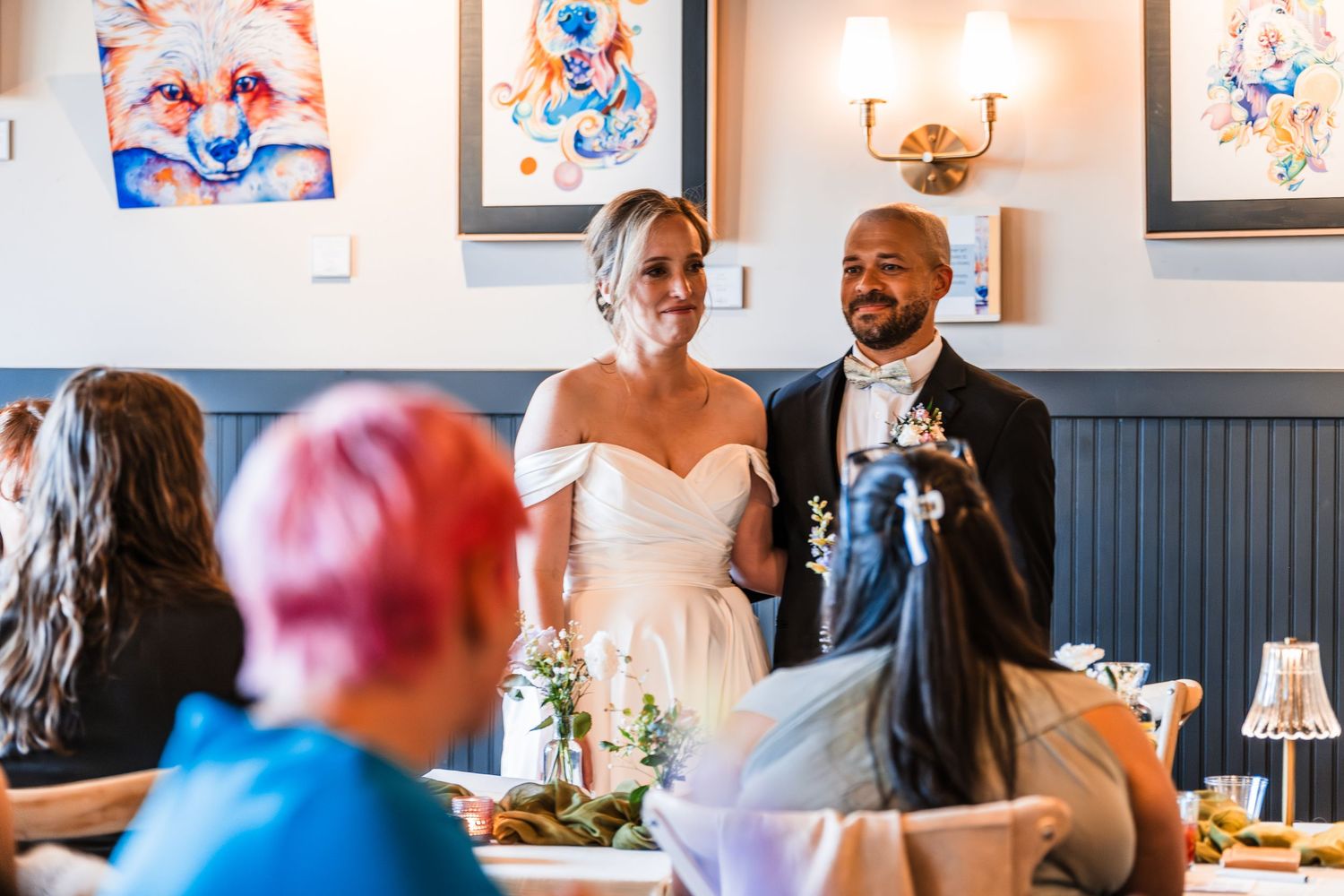 Bride in off-shoulder gown and groom with boutonniere smiling at guests during wedding reception.